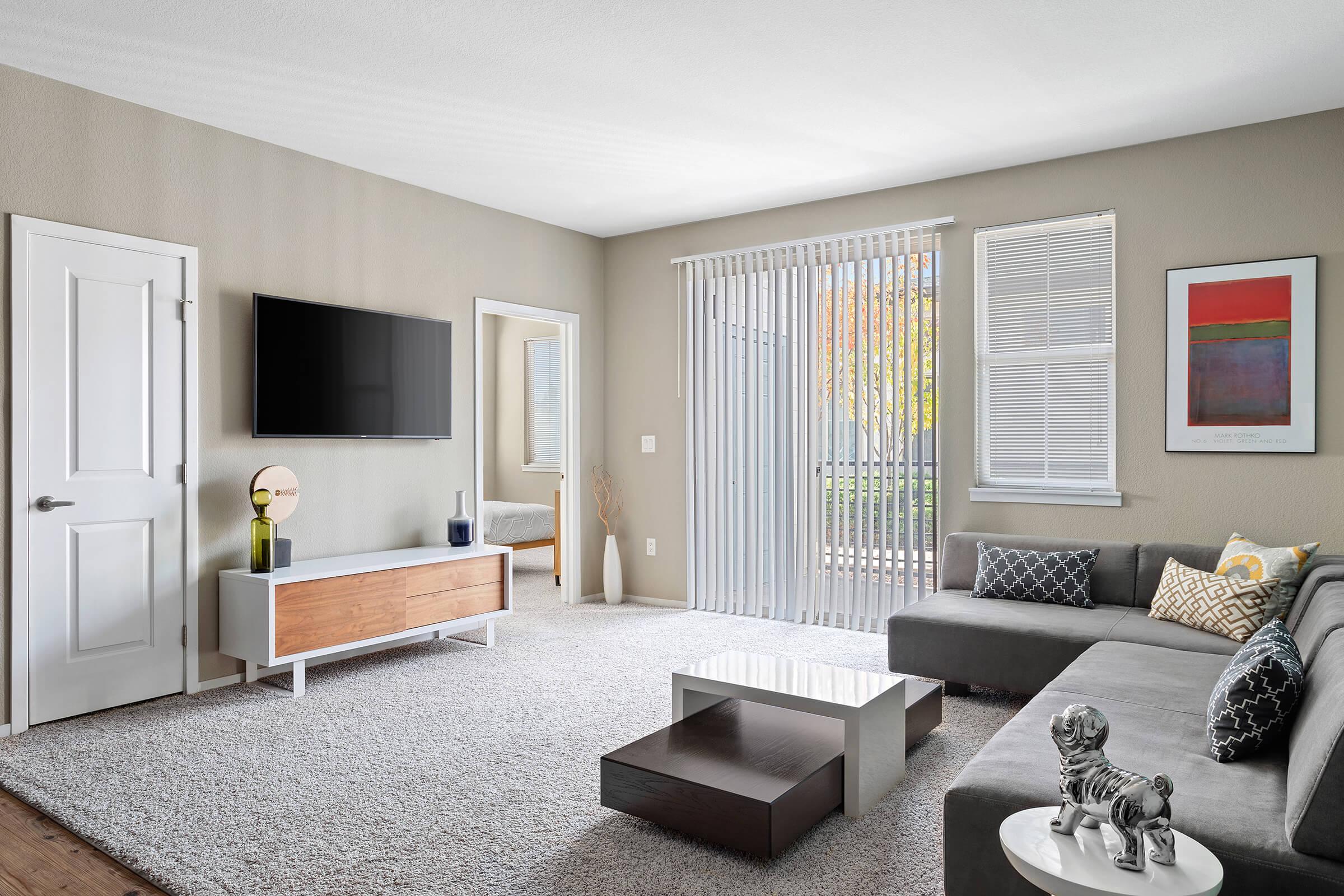 A modern living room featuring a gray sectional sofa with decorative pillows, a coffee table, a flat-screen TV mounted on the wall, and a stylish wooden media console. Large windows with vertical blinds allow natural light in, and a piece of artwork hangs on the wall.