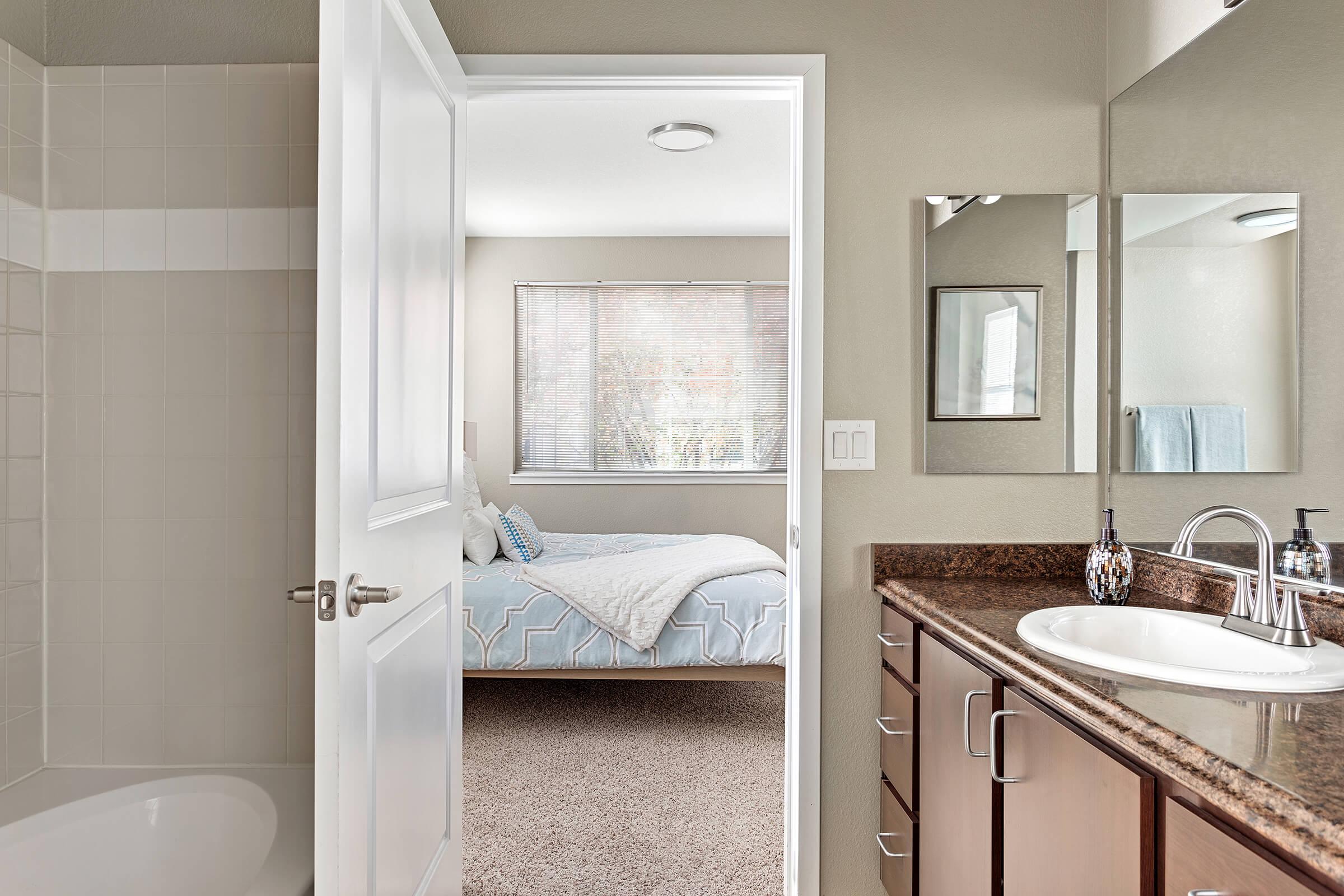 A view of a bathroom doorway leading to a bedroom. The bathroom features a shower tub combo, a sink with a granite countertop, and a large mirror. The adjoining bedroom has a bed with light bedding and a large window with natural light filtering in. The overall color scheme is neutral and inviting.