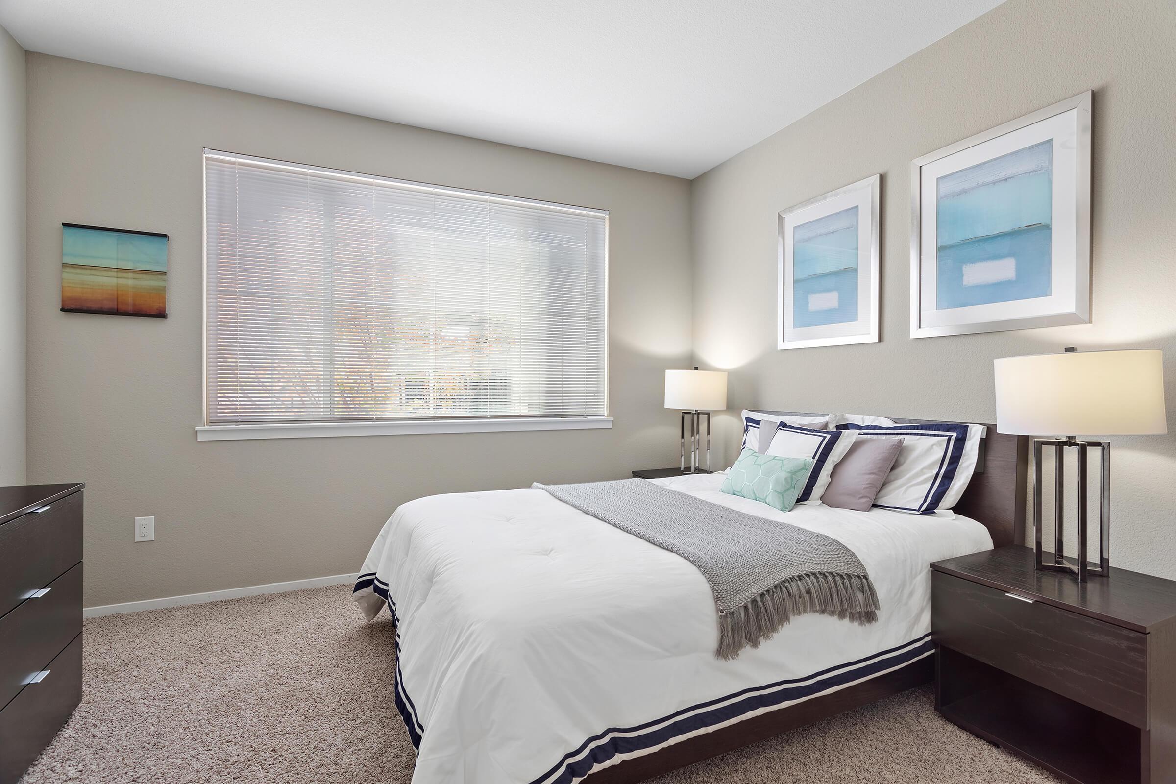 Cozy bedroom featuring a bed with white bedding, a light gray throw blanket, and decorative pillows. Two bedside tables with lamps are on either side of the bed. A window with blinds allows natural light to illuminate the room, which has soft gray walls and framed artwork above the bed.