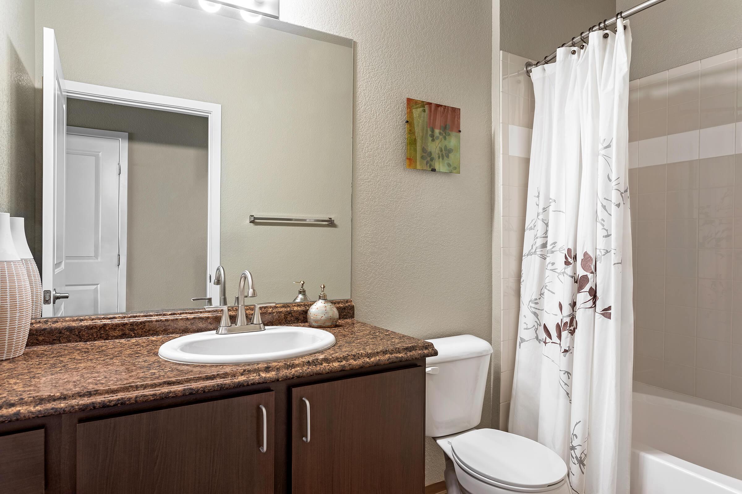 A neatly designed bathroom featuring a brown granite countertop with a sink, a white toilet, and a shower with a white curtain adorned with floral patterns. The walls are painted a soft green, and there is a mirror above the sink along with a small piece of art on one wall.