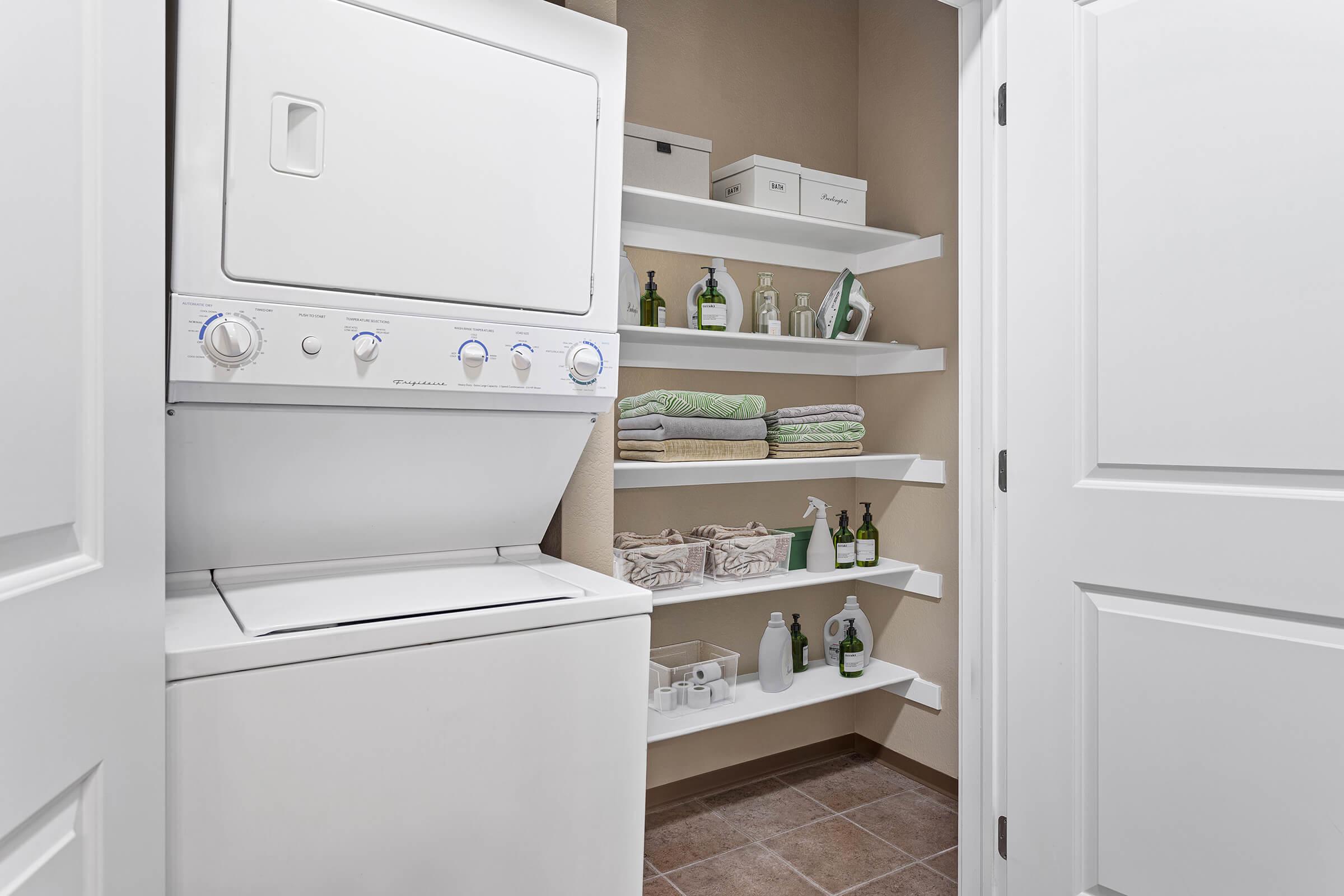 A compact laundry area featuring a stacked washer and dryer, white shelving displaying neatly arranged cleaning supplies, bottles, and folded towels in soft green and white colors. The walls are painted a neutral tone, creating a clean and organized space.