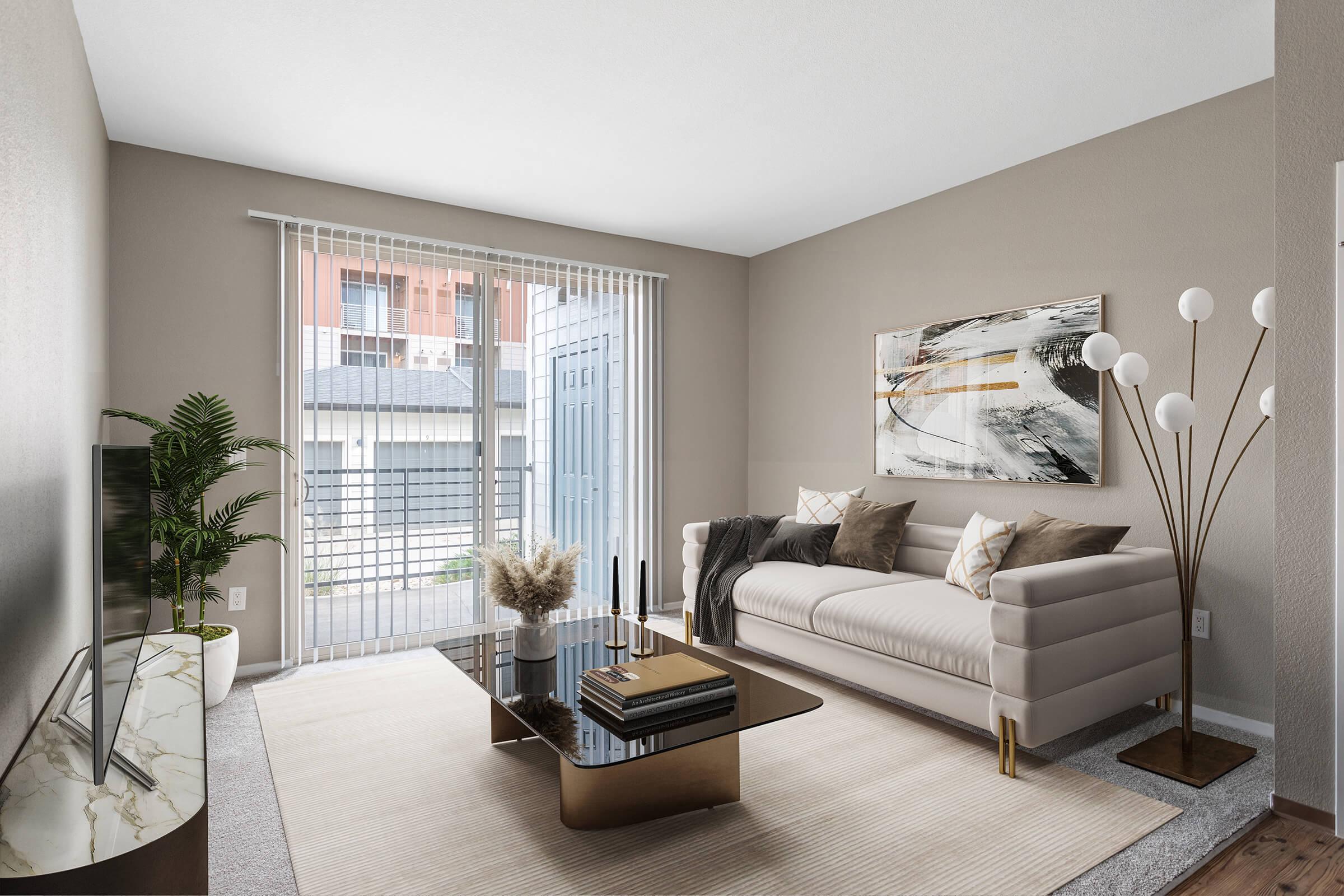 A spacious, empty room with light beige walls and carpet. Large sliding glass doors with vertical blinds lead to a balcony. The room appears bright and airy, with a minimalist design and no furniture. Natural light enters from outside, providing a welcoming atmosphere.