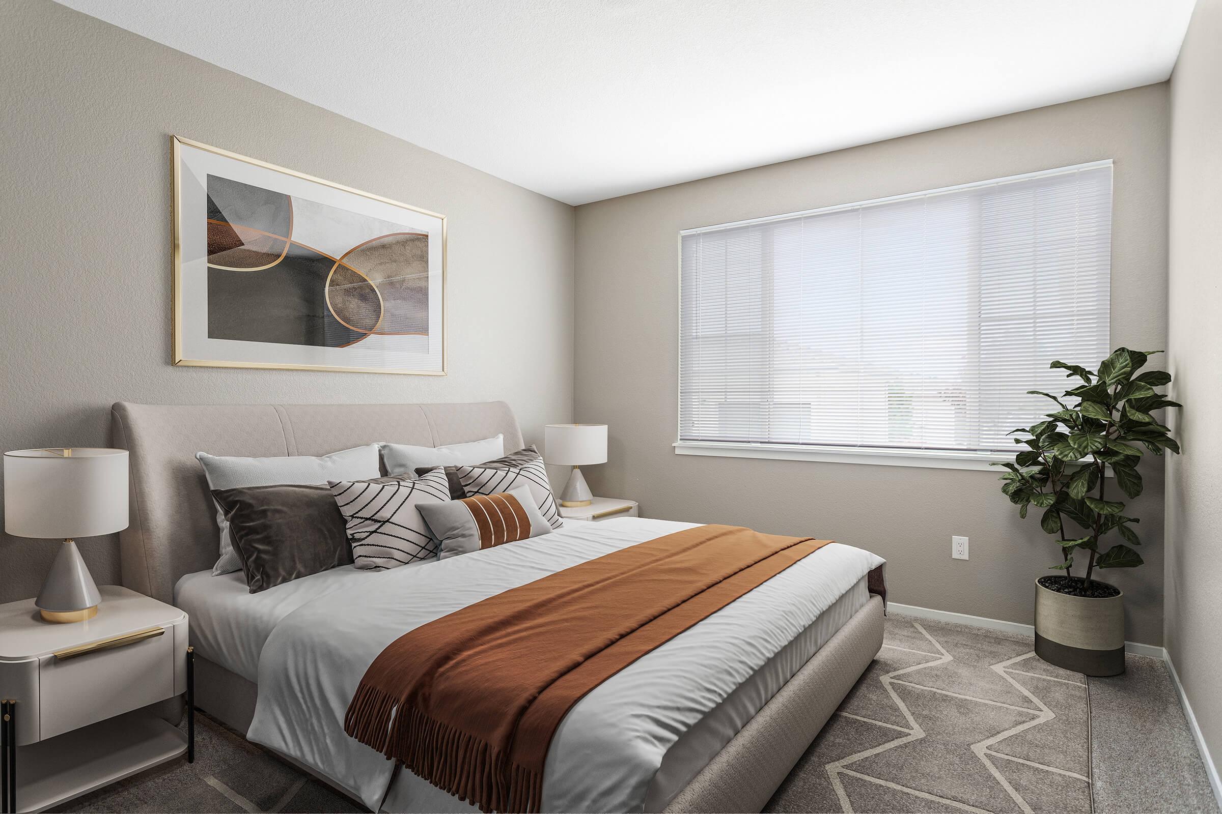 A cozy bedroom featuring a queen-sized bed with a patterned throw blanket, accented by two bedside lamps and a framed abstract artwork on the wall. A lush green plant adds a touch of nature, while natural light streams through a large window with blinds, creating a warm and inviting atmosphere.