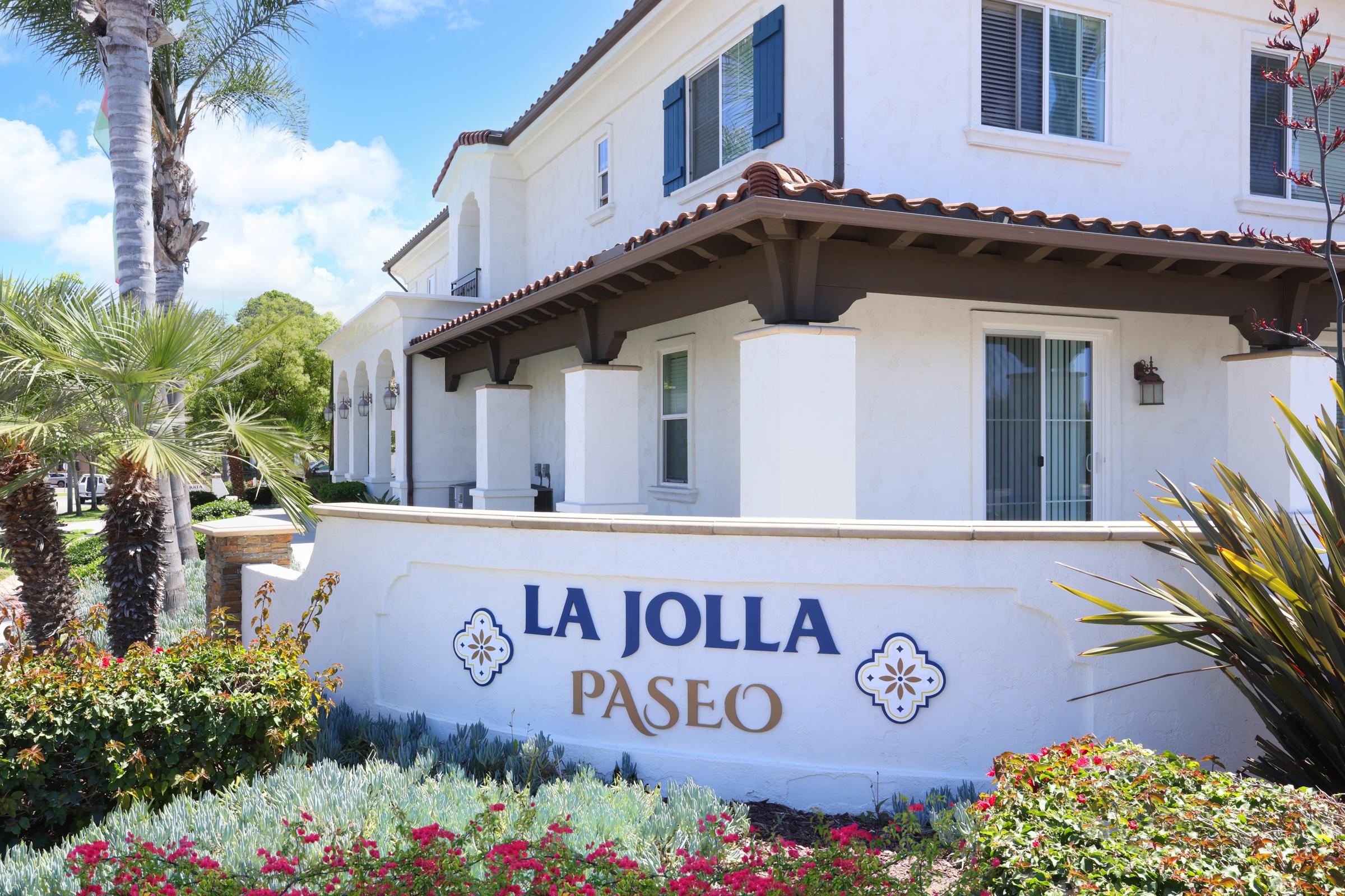 A view of a white building with a tiled roof, featuring the words "La Jolla Paseo" prominently displayed. Surrounding the building are green plants and colorful flowers, with palm trees in the background under a blue sky.