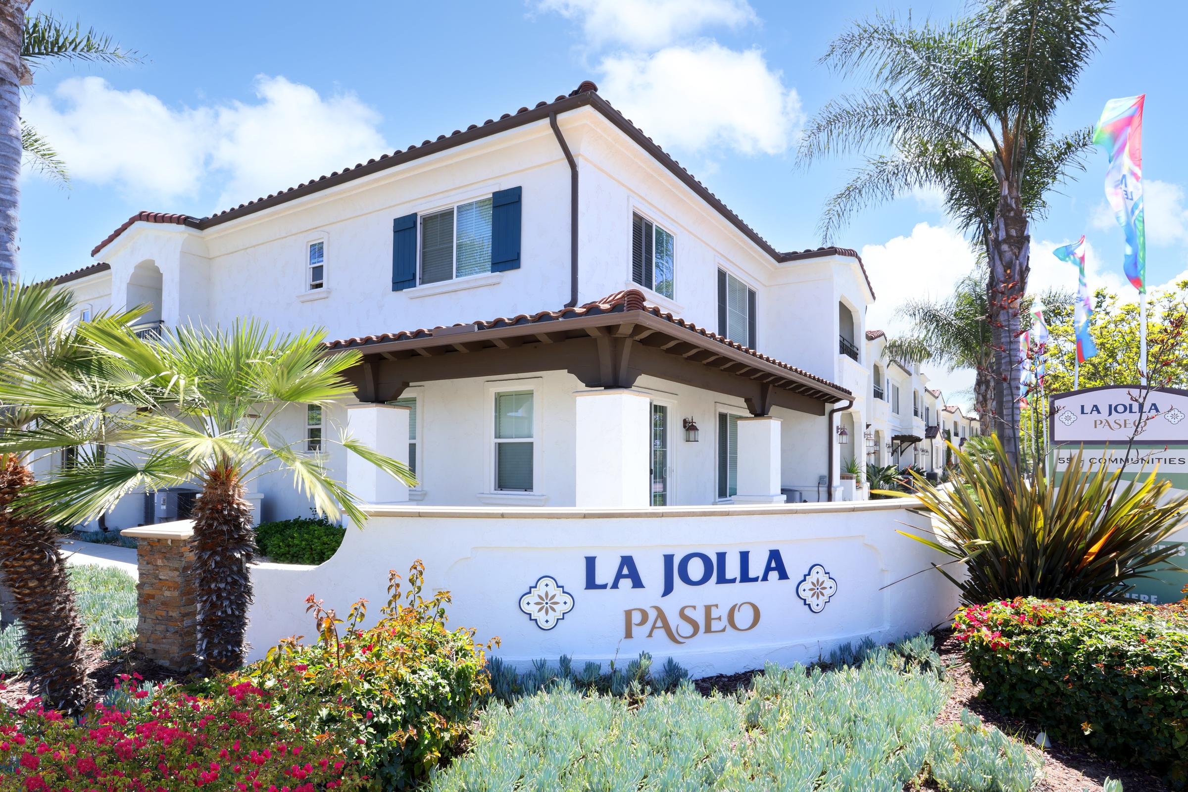 A white stucco building with tiled roofs, featuring green shutters and palm trees in the foreground. The sign reads "LA JOLLA PASEO," indicating it's a residential or commercial complex. The sky is blue with a few clouds, creating a bright and inviting atmosphere.