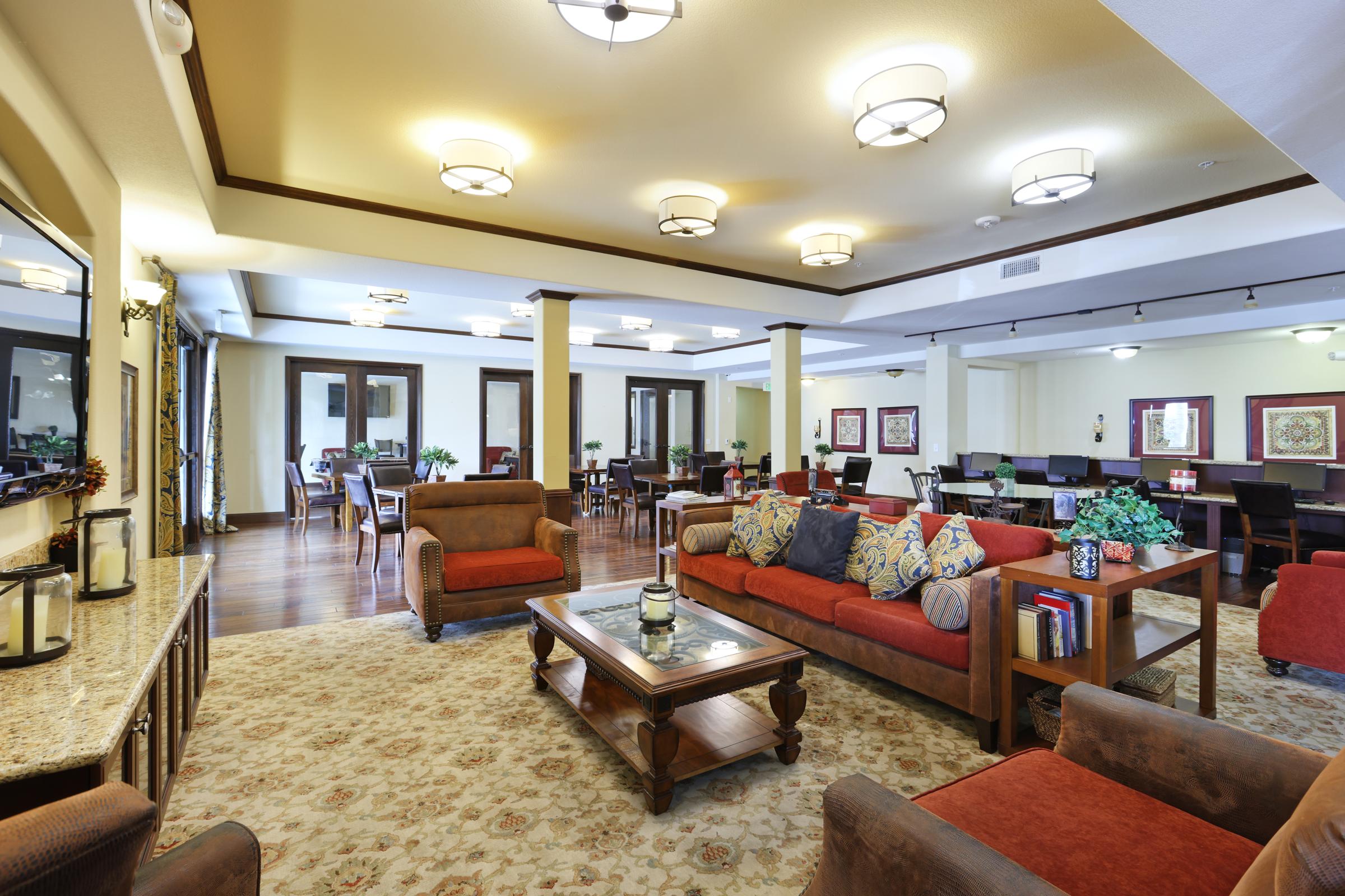 A spacious and inviting common area featuring comfortable seating, decorative pillows, and a central coffee table. The space has soft lighting, wooden accents, and a cozy atmosphere, with dining tables in the background and floral artwork on the walls. A warm color palette enhances the welcoming ambiance.