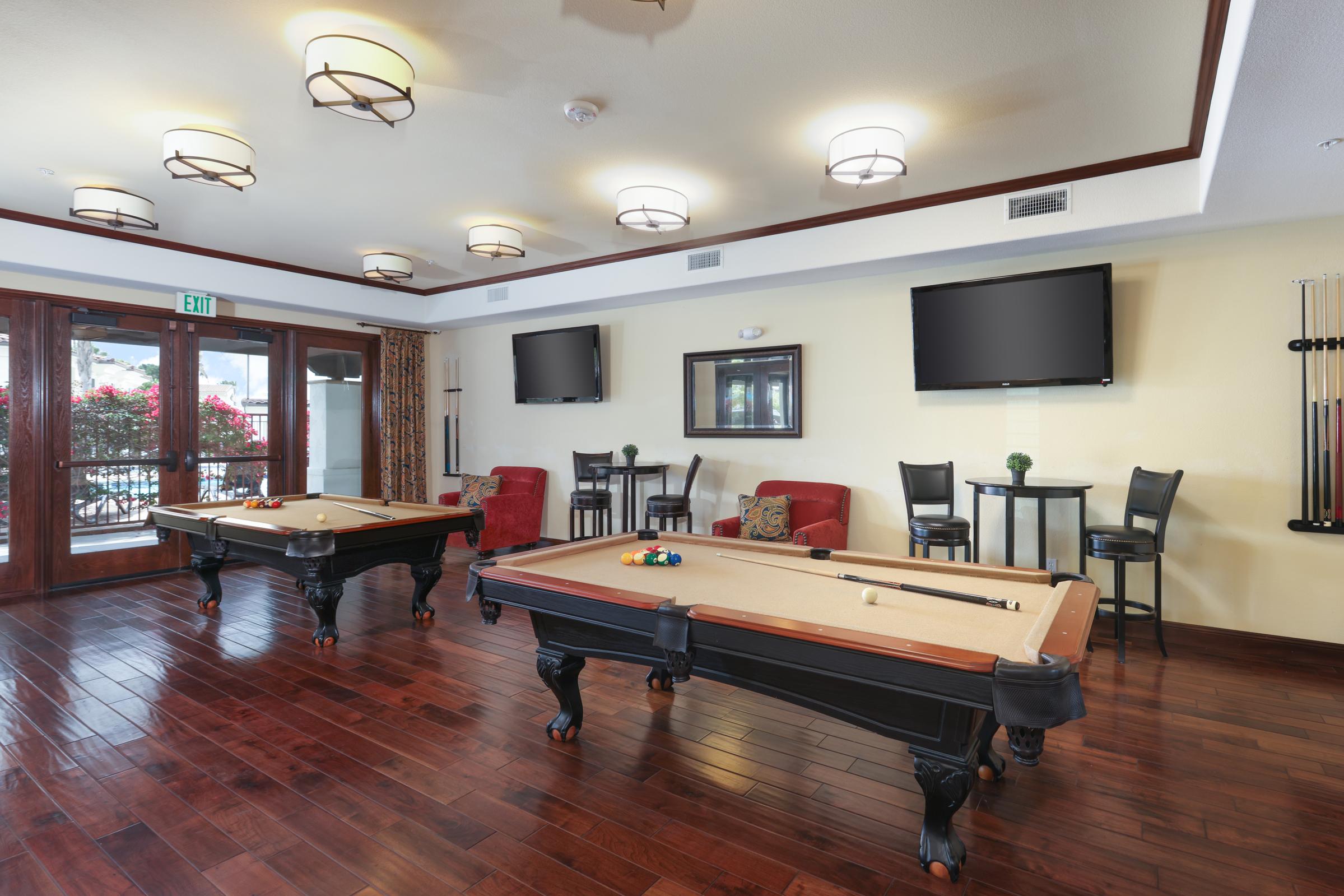 Interior view of a recreational room featuring two pool tables, wooden flooring, and stylish lighting. There are seating areas with red chairs and small tables, and two flat-screen TVs mounted on the walls. Large windows allow natural light and views of outdoor greenery.