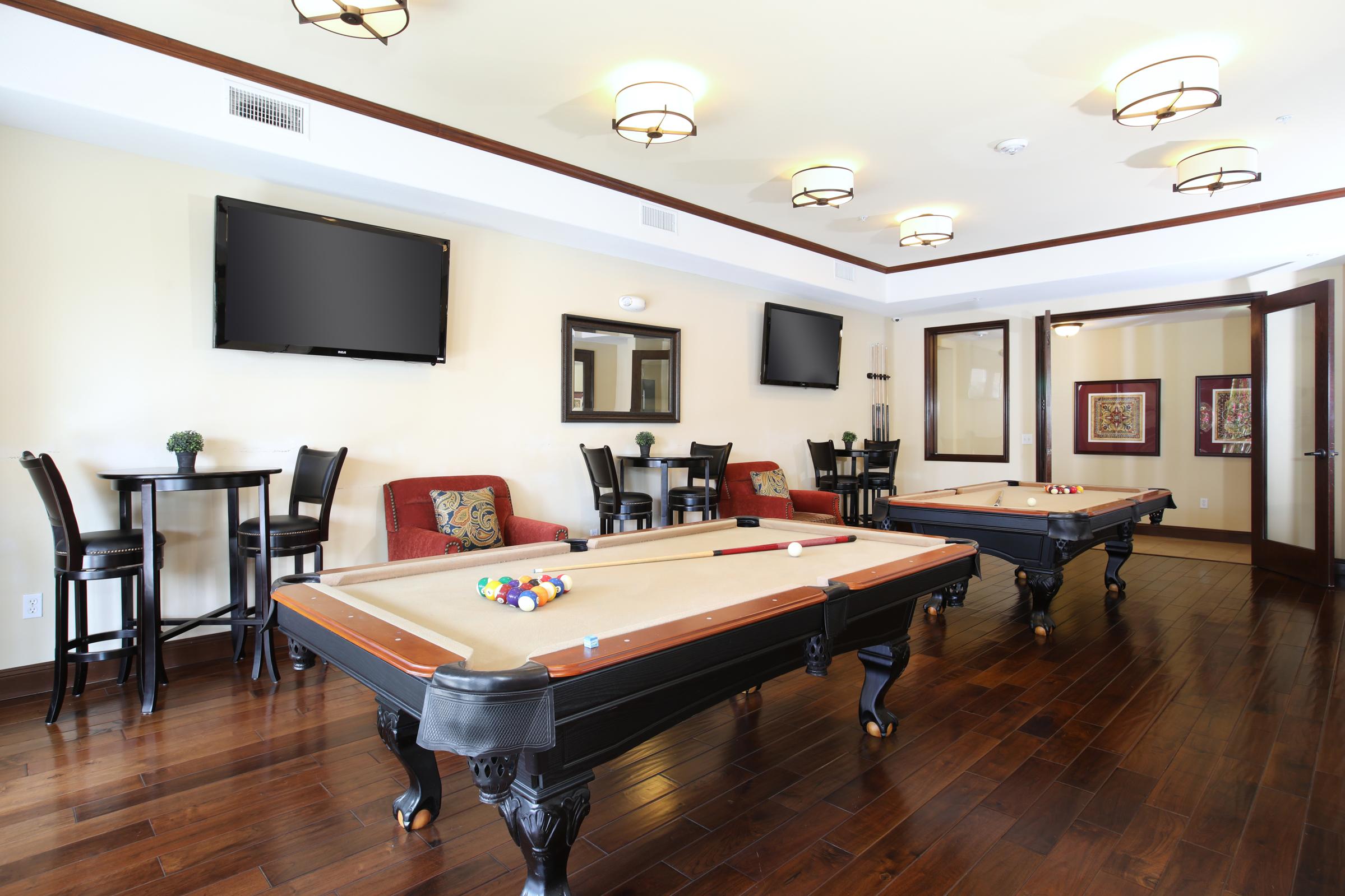 A stylish common area featuring two pool tables, several seating options with plush chairs, and wall-mounted televisions. The walls are painted in a light color, adorned with decorative mirrors and artwork, while the hardwood floor adds a warm touch to the inviting atmosphere.