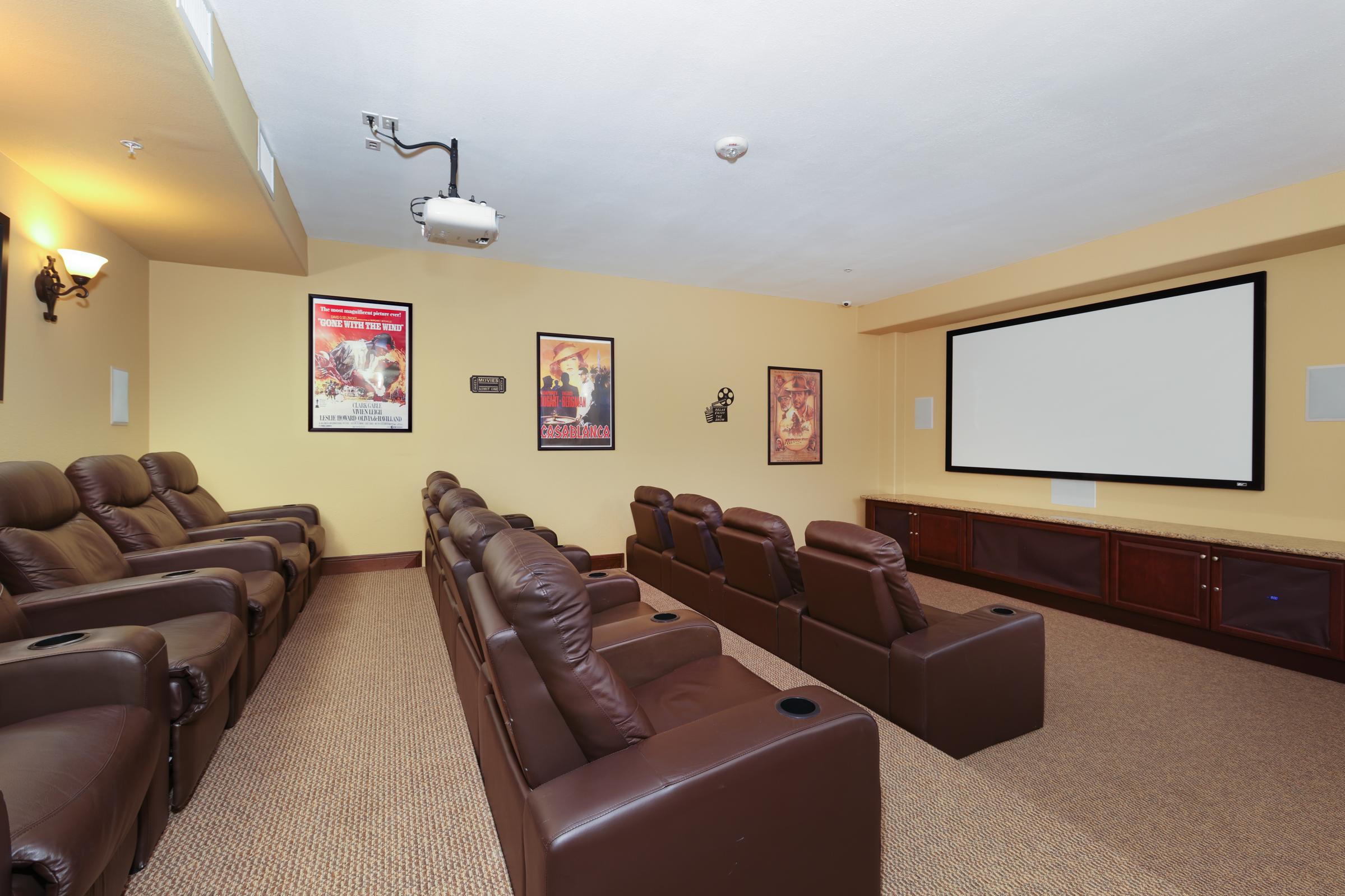 Home theater setup featuring plush, reclining leather chairs arranged in rows facing a large projection screen. The walls are decorated with movie posters, and there is a projector mounted on the ceiling. Warm lighting creates a cozy atmosphere in the room.