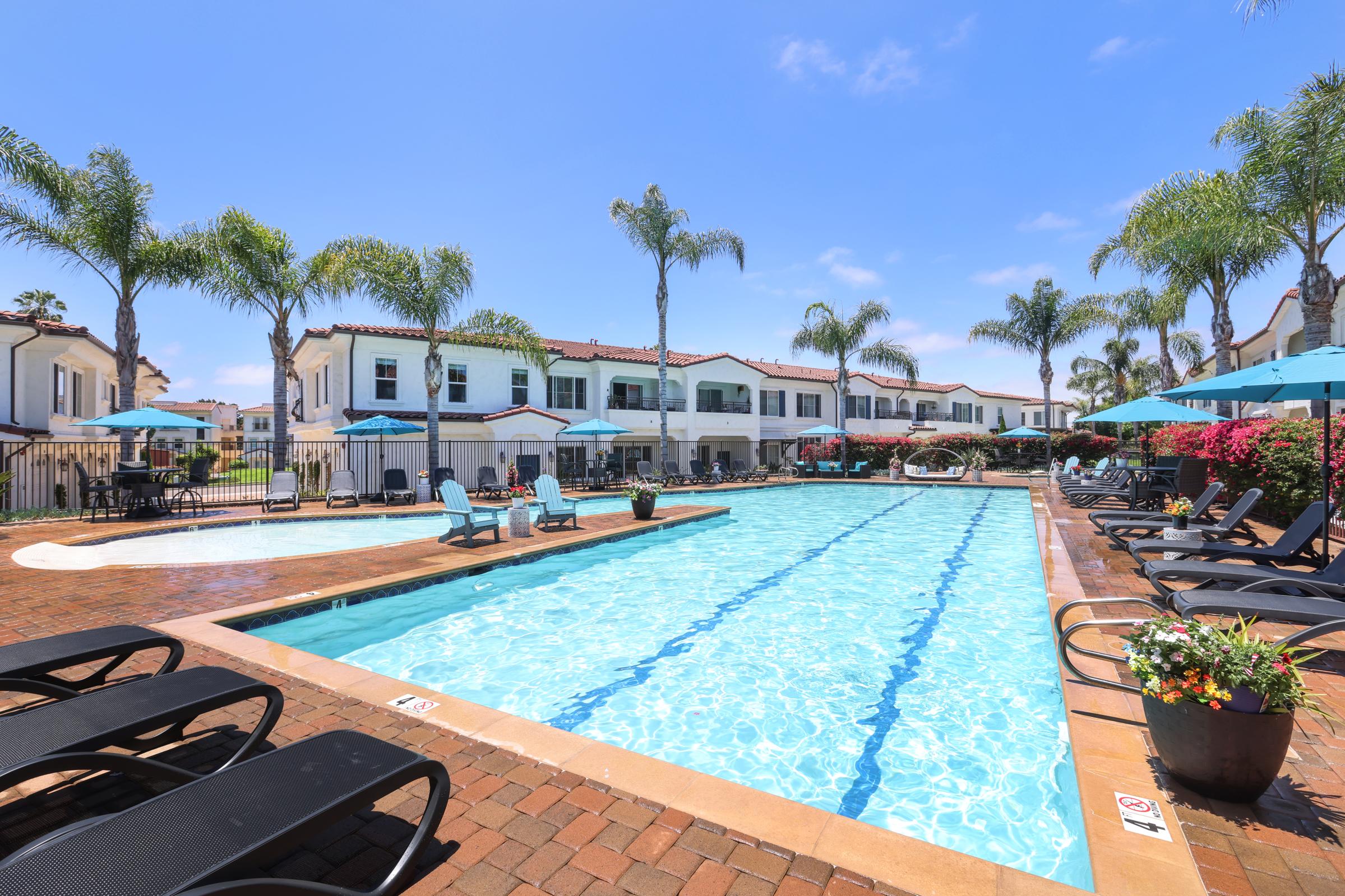 A refreshing swimming pool surrounded by palm trees and lounge chairs. Bright blue skies overhead and comfortable seating with umbrellas create a relaxing atmosphere. Nearby, elegant buildings add to the serene setting, with vibrant flowers enhancing the landscape. Perfect for leisure and relaxation.