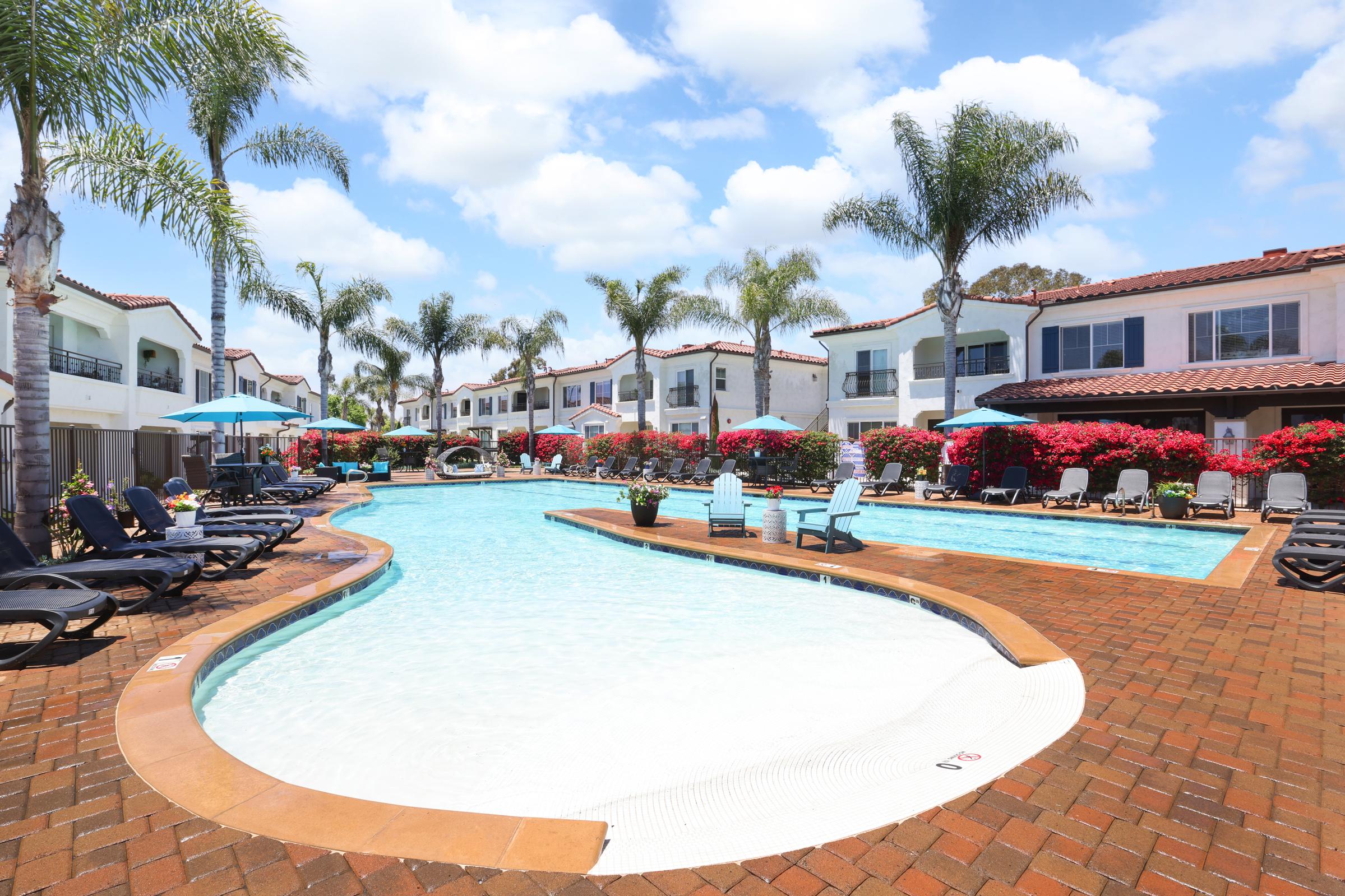 A vibrant swimming pool area surrounded by palm trees and colorful flowers. Lounge chairs line the poolside, and umbrellas provide shade. Bright blue skies with fluffy clouds overhead create a relaxing atmosphere in this outdoor leisure space.