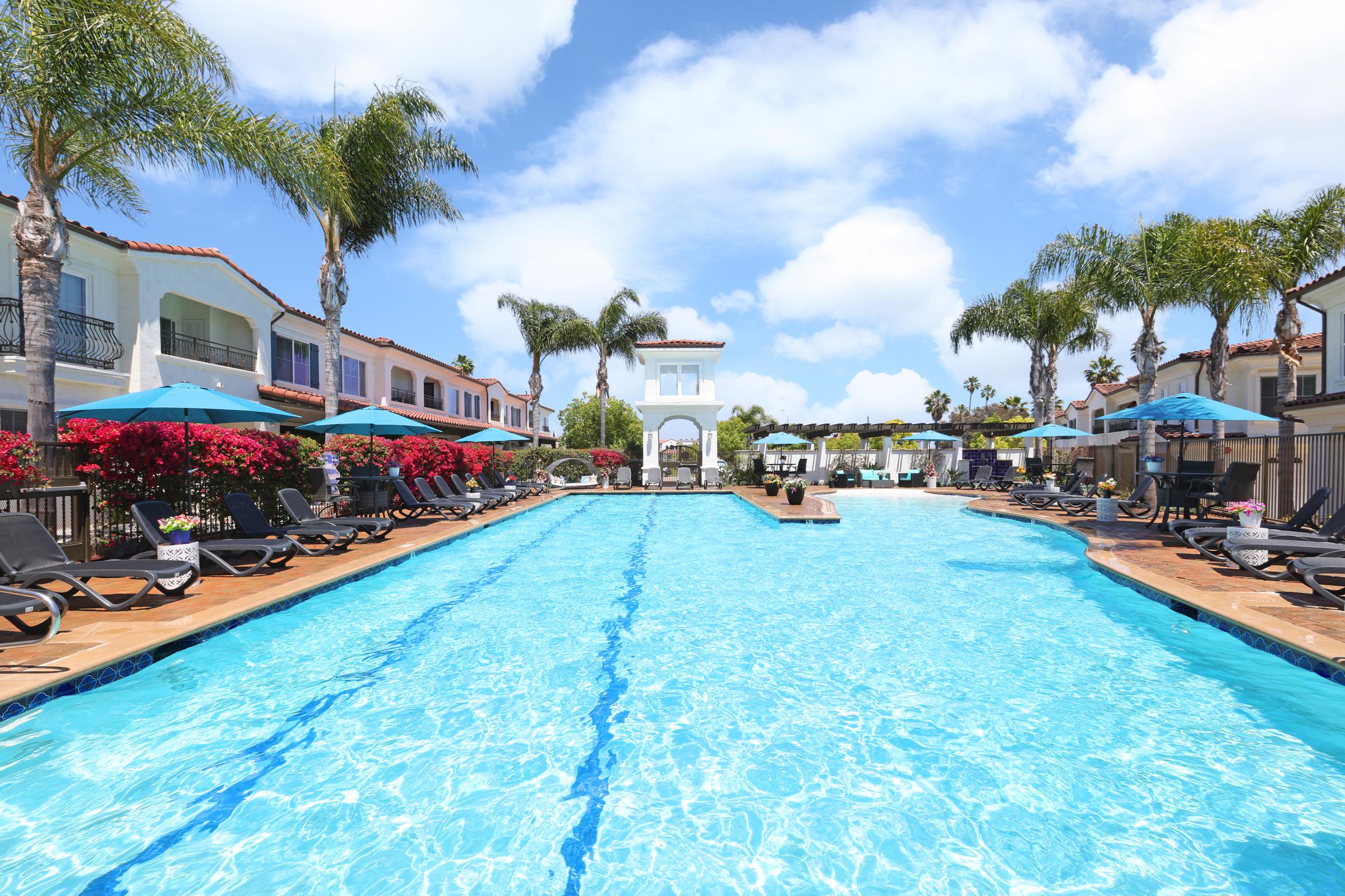 A vibrant outdoor pool area featuring a clear blue pool surrounded by lounge chairs and large umbrellas. Lush palm trees and colorful flower beds add to the tropical atmosphere. Bright blue skies with scattered clouds create a cheerful ambiance, ideal for relaxation.