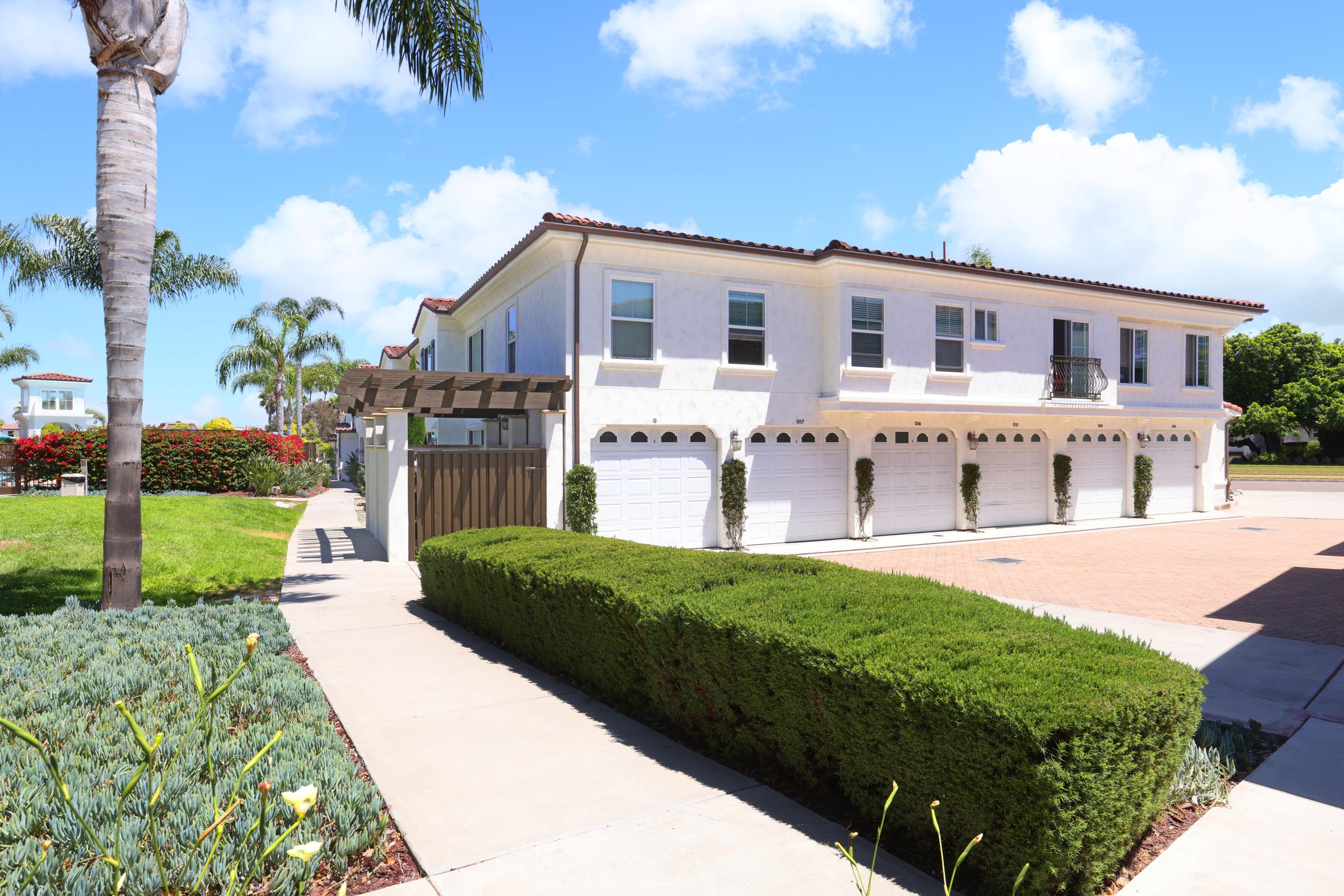 A modern two-story building with a white exterior and multiple garage doors. The surrounding area features well-maintained landscaping, including green hedges and palm trees, under a bright blue sky with fluffy white clouds.