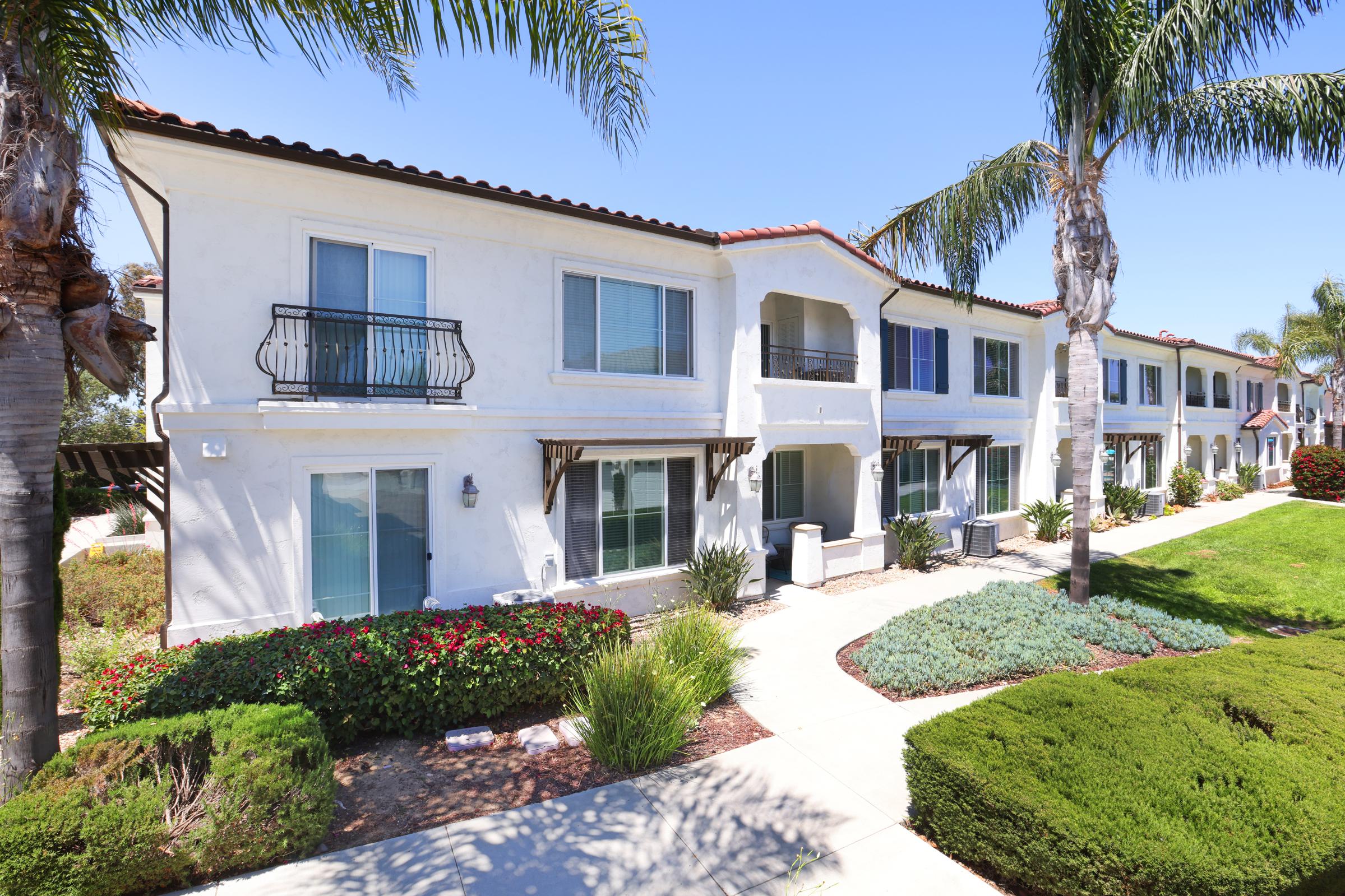 A row of elegant two-story residential buildings with white stucco exteriors. Each unit features large windows, decorative balconies, and landscaped grounds with green shrubs and palm trees. The pathway leads through a well-maintained garden area, under clear blue skies.