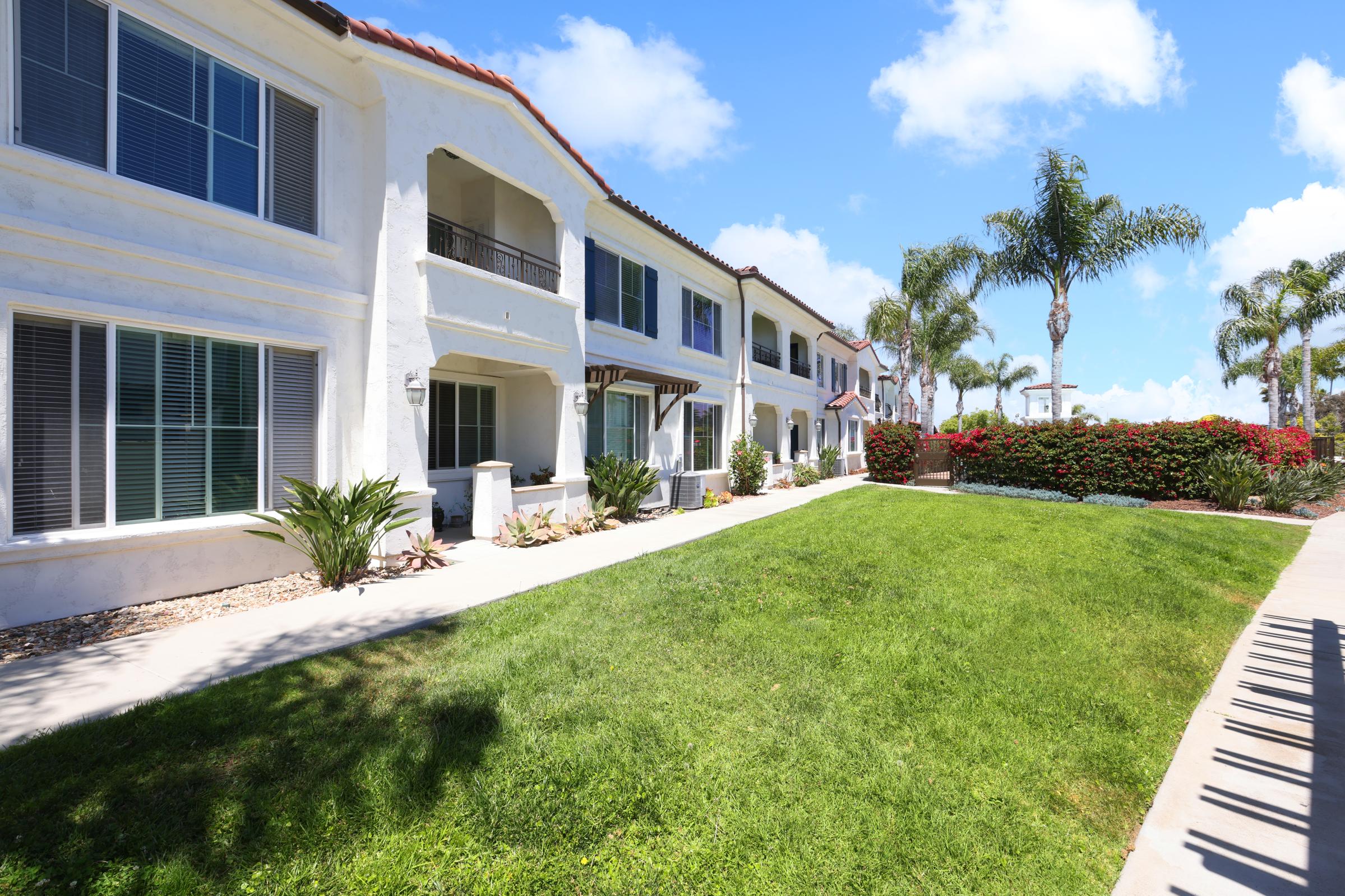 A well-maintained residential area featuring two-story white buildings with balconies, surrounded by lush green grass and decorative plants. Palm trees and vibrant flower beds provide a tropical feel under a clear blue sky. The setting exudes a serene and inviting atmosphere.