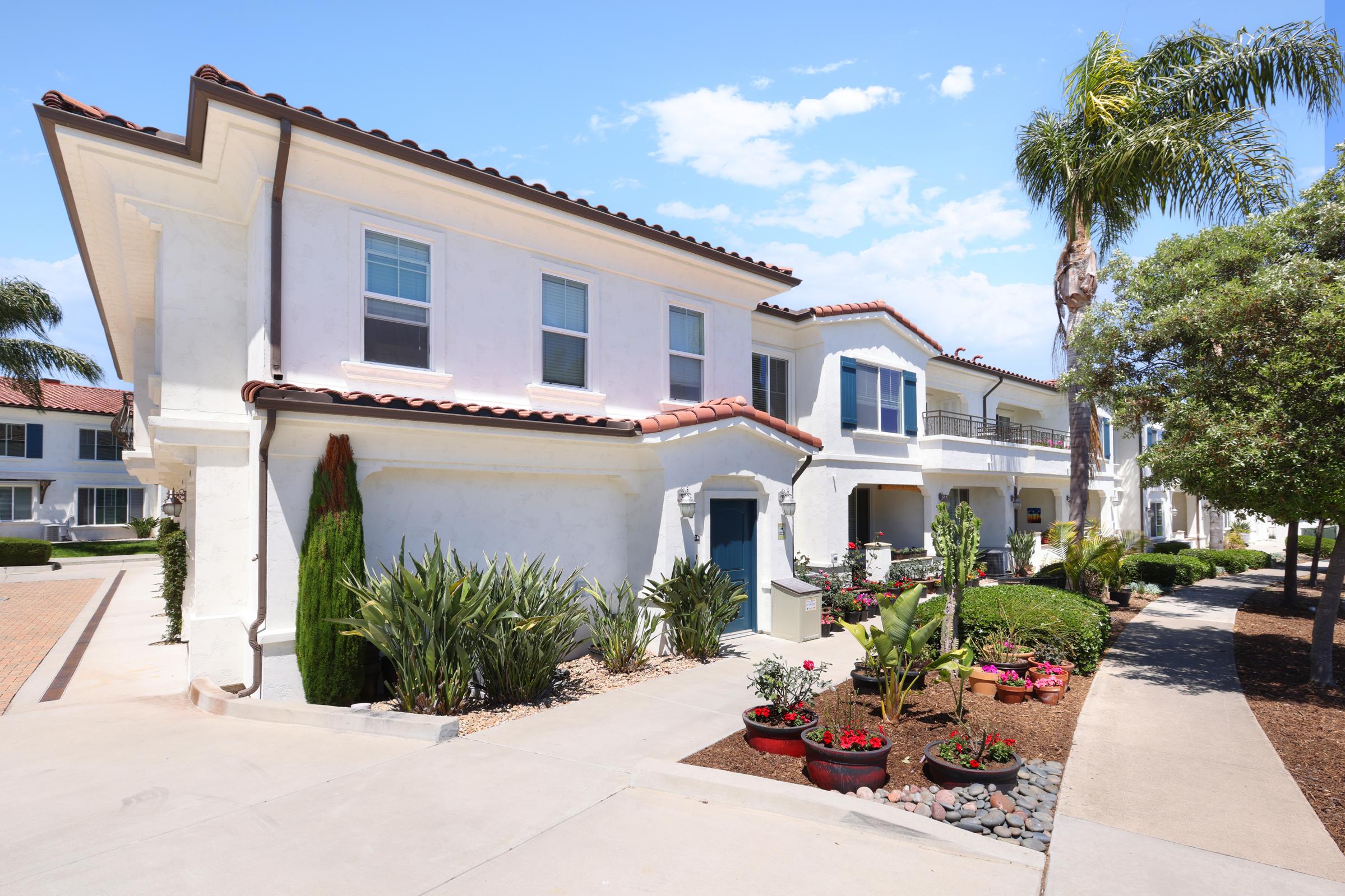 A sunny residential area featuring white stucco buildings with red-tiled roofs. The buildings are surrounded by lush green landscaping, including palm trees, shrubs, and flower beds with colorful plants. A paved pathway leads to the front entrances of the units. Clear blue skies above.