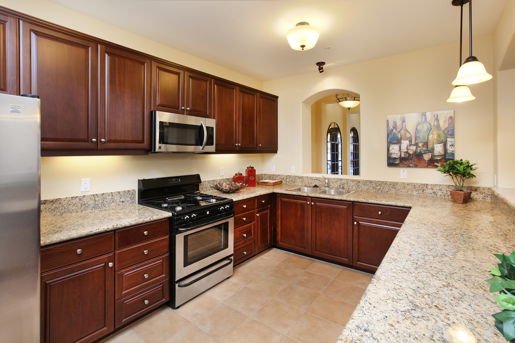 A modern kitchen featuring dark wood cabinetry, stainless steel appliances, and a granite countertop. The kitchen includes a gas stove, microwave, and sink, with tile flooring and warm lighting fixtures. Decorative elements like a framed artwork and a potted plant add a homey touch.