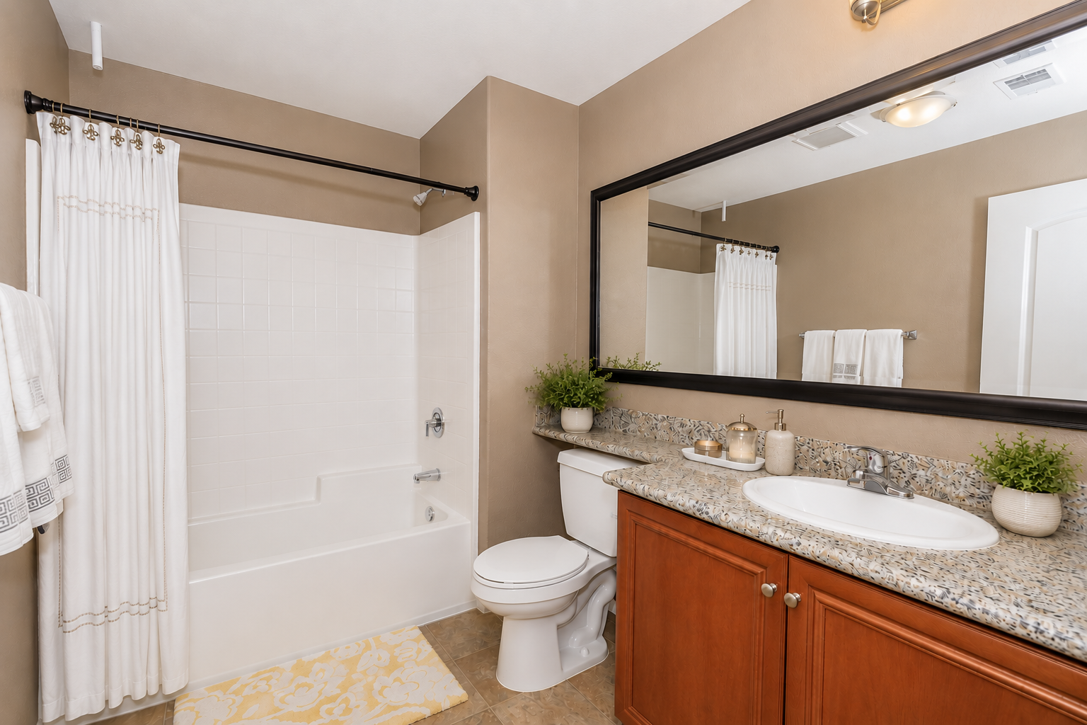 A modern bathroom featuring a shower-tub combo, beige walls, a large mirror above a sink with a granite countertop. There's a white toilet, decorative plants, and towels neatly arranged. Natural light brightens the space, giving it a clean and inviting look.