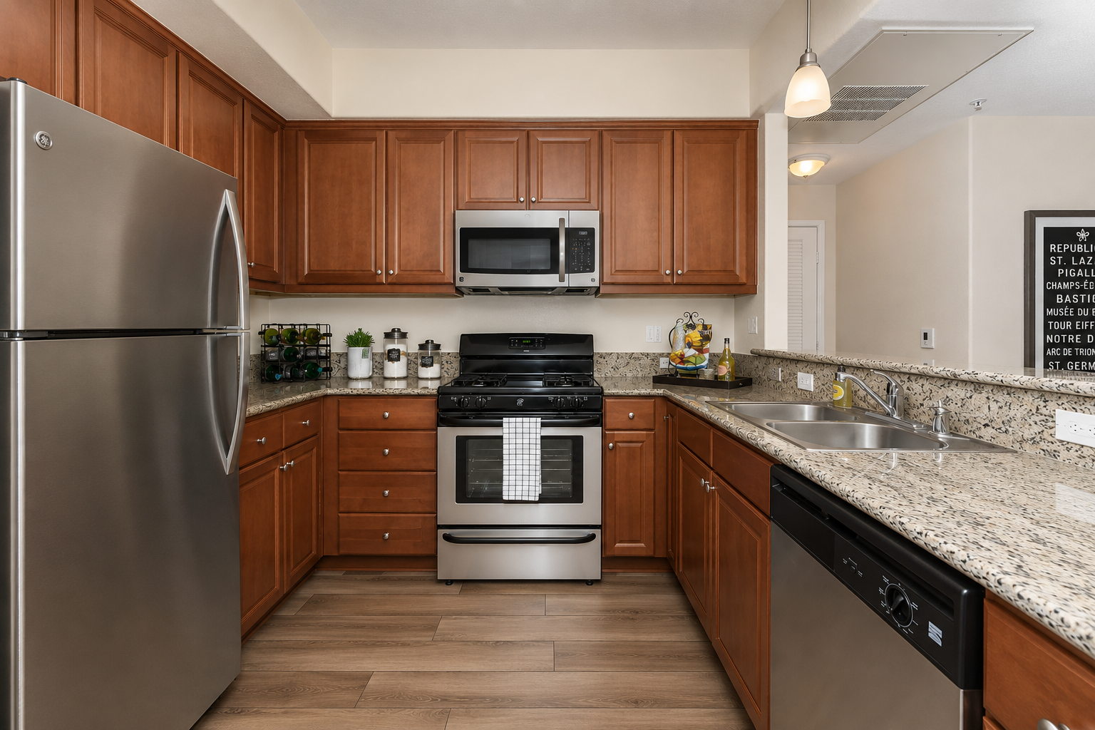 Modern kitchen featuring wood cabinetry, stainless steel appliances including a refrigerator, oven, microwave, and dishwasher. The countertop is made of granite, and there's a double sink with a sleek faucet. Decorative plants and a bowl of fruit add a touch of color to the space.