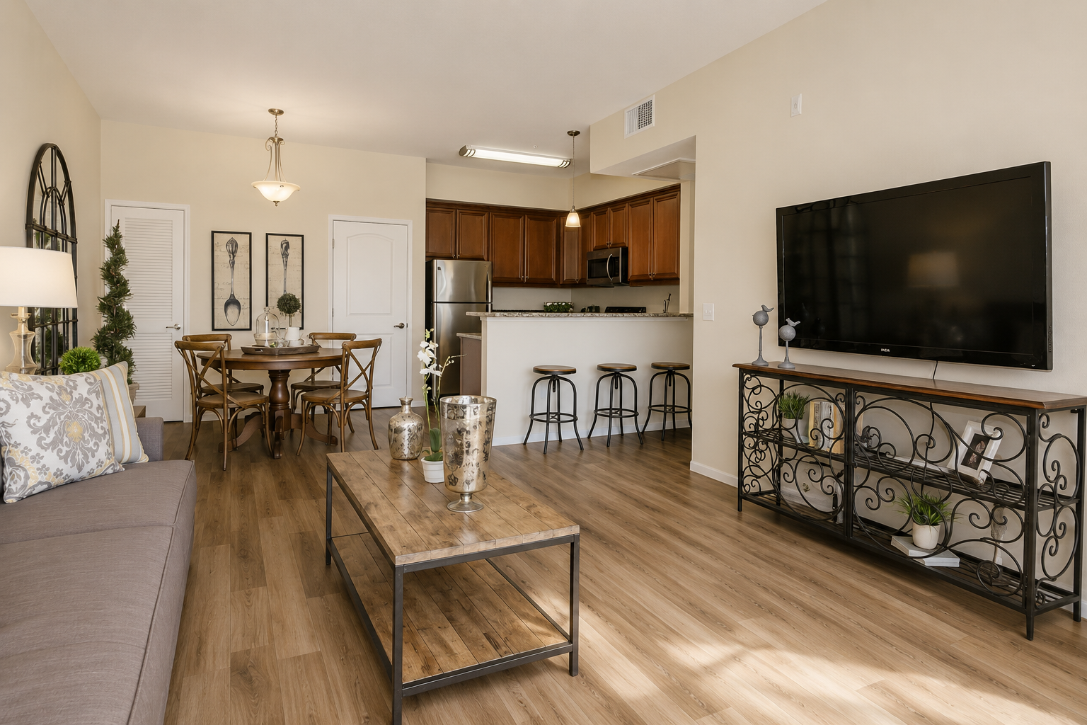 A modern living room featuring a gray sofa, a wooden coffee table with decorative vases, and a TV stand holding a flat-screen TV. In the background, there's a dining area with a table and chairs, and a kitchen with wooden cabinets. The space is well-lit and decorated with plants.