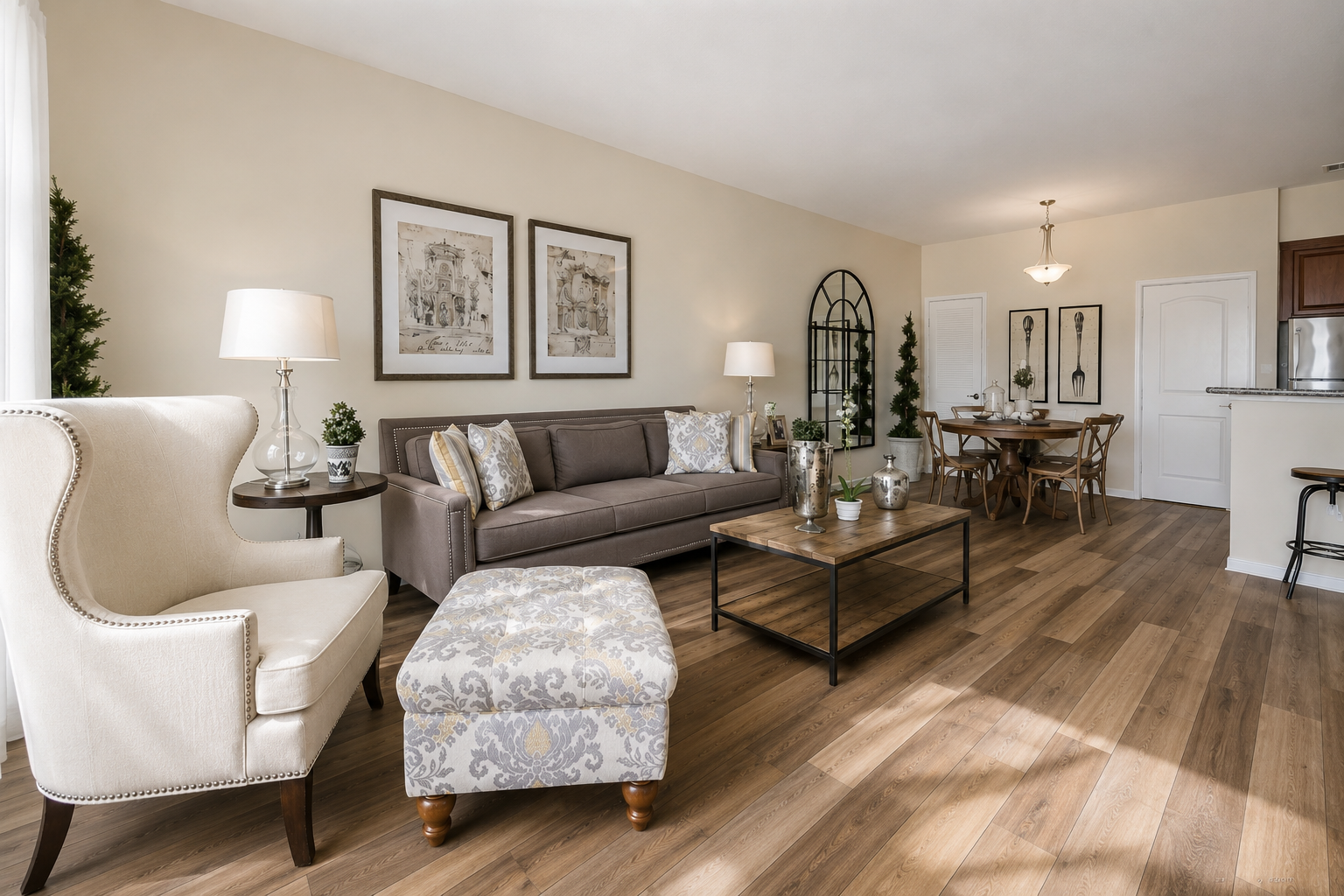 A stylish living room featuring a beige wall, a comfortable brown sofa with decorative pillows, a patterned ottoman, and a coffee table. The room is illuminated by natural light, and there are framed artworks on the wall. In the background, there's a dining area with a wooden table and chairs.