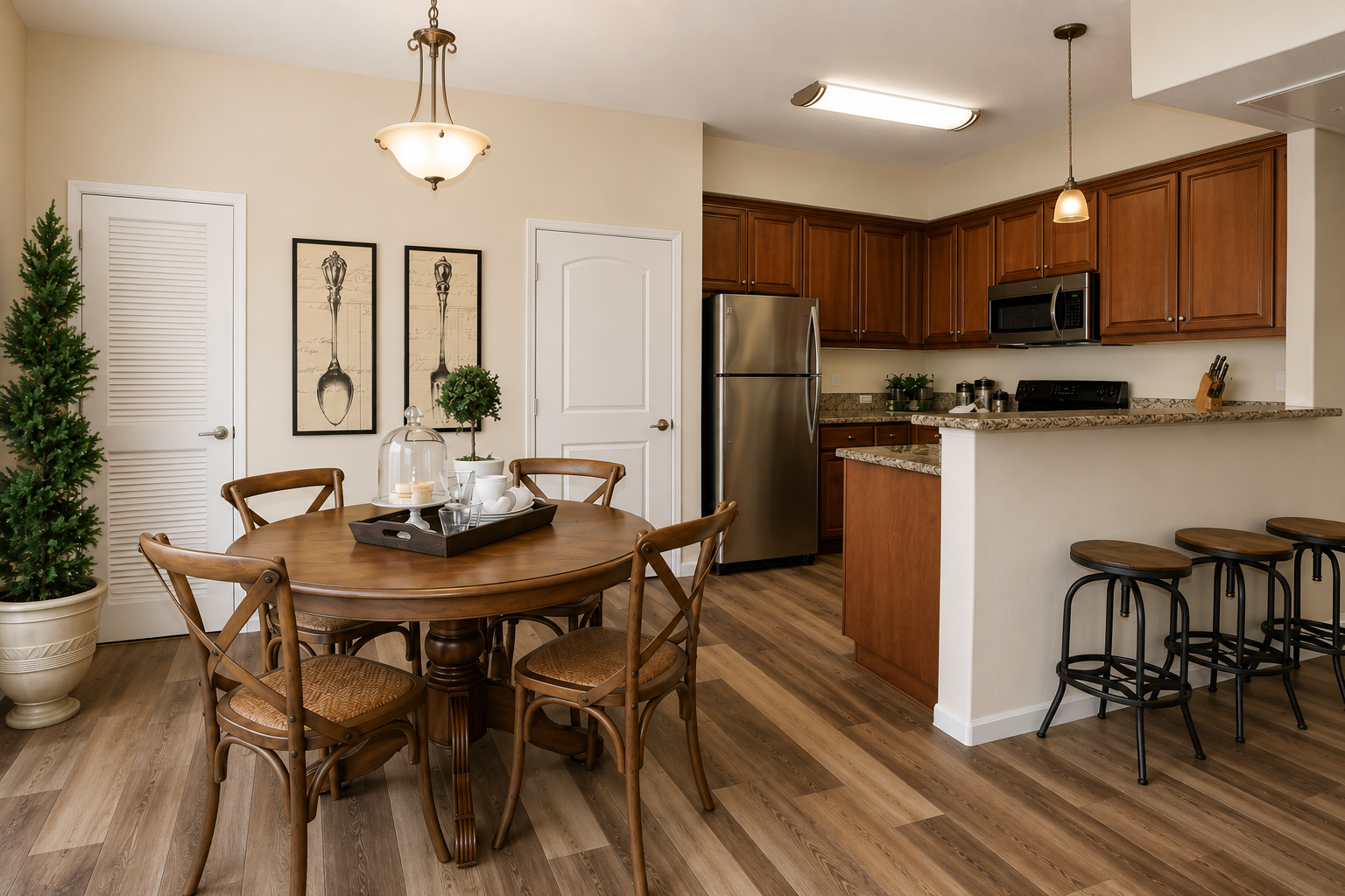 A modern kitchen and dining area featuring a round wooden table with four chairs, a decorative centerpiece, and a bar counter with stools. The kitchen includes dark wood cabinets, stainless steel appliances, and a light-colored countertop. The room is bright and warm with light walls and wooden flooring.