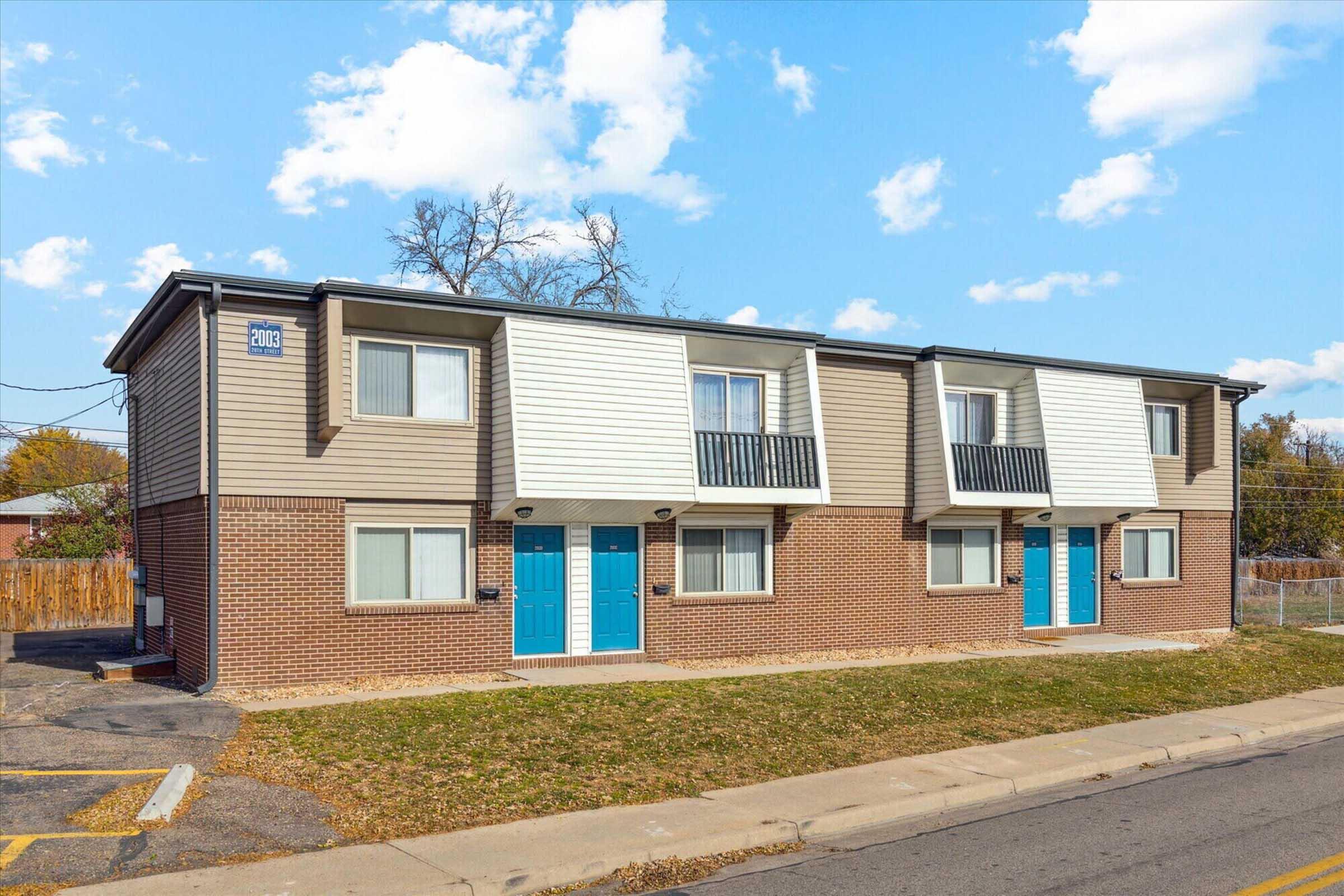 Exterior view of a two-story, multi-unit residential building featuring modern architecture. The building has brick and siding exteriors with large windows and blue doors. Surrounding landscaping includes grass and a paved walkway. The sky is partly cloudy.