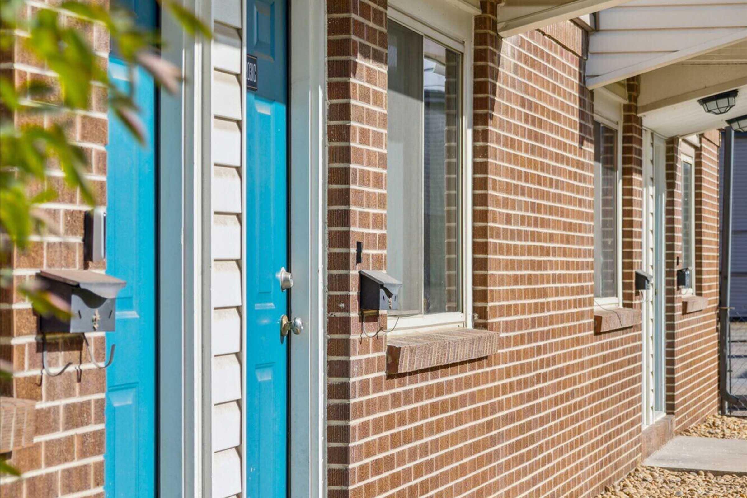 Two blue doors with silver handles are set into a brick wall of a building. The brick has a warm reddish-brown hue. There are small mailboxes mounted next to each door. A green plant is partially visible on the left side, adding a touch of color to the scene. The setting appears to be a well-maintained residential area.