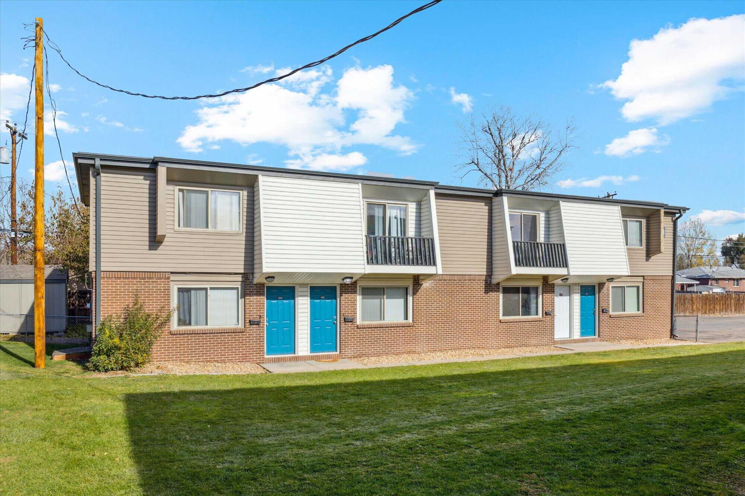 Two-story apartment building with a light brown exterior and white panels, featuring balconies. The front has two blue doors on each unit, surrounded by a well-maintained lawn and a clear blue sky with some clouds in the background. Power lines run alongside the building.