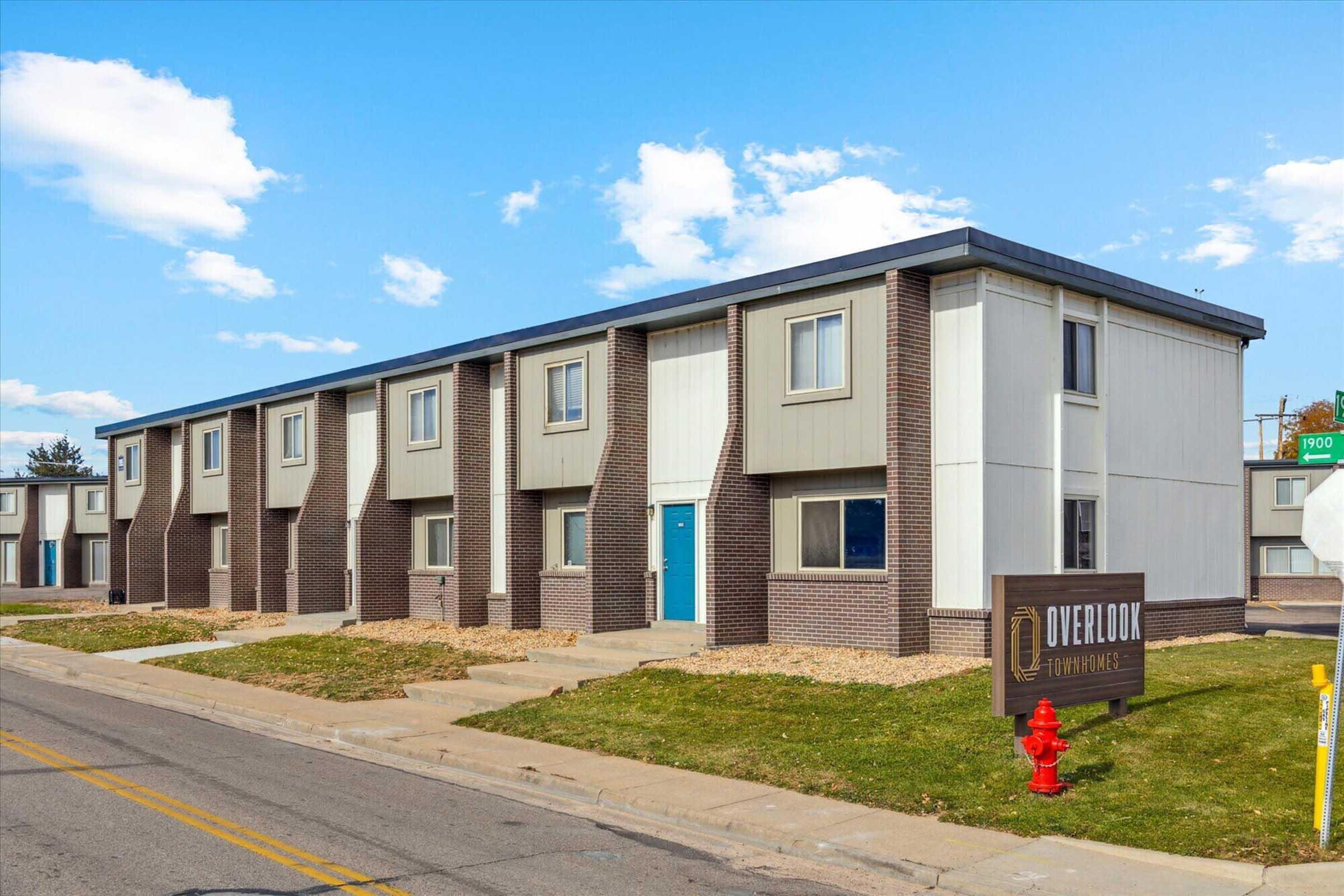 A low-rise apartment building with a modern exterior featuring a combination of brick and siding. The building has multiple windows and a blue door. In front, there is a sign that reads "Overlook Apartments," and a red fire hydrant near the sidewalk. The scene is set against a clear blue sky.