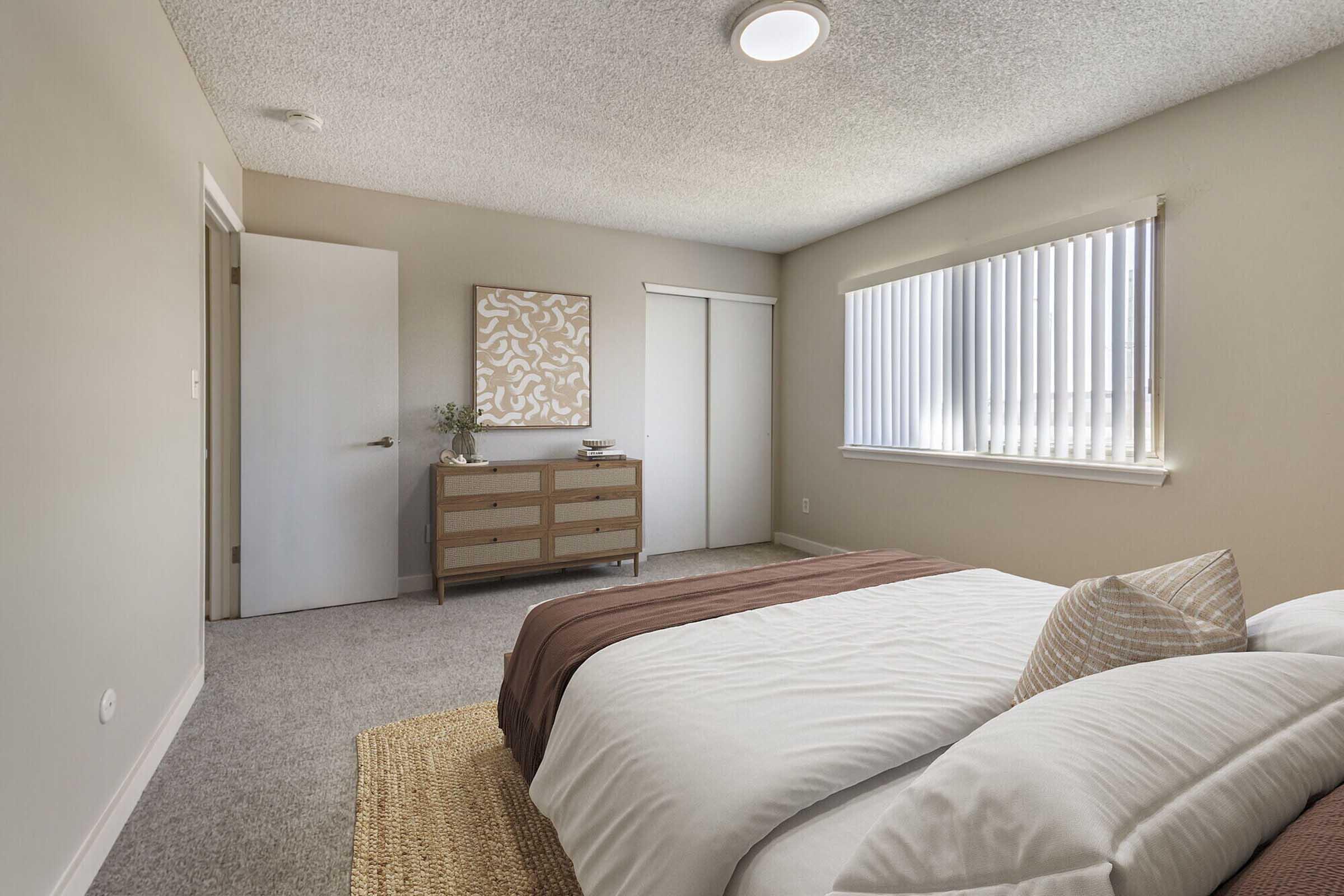 A cozy bedroom featuring a neatly made bed with brown and white bedding, a textured pillow, and a woven rug. There is a wooden dresser with decorative items, a large window with vertical blinds, and a door leading to a closet. The walls are painted in neutral tones, creating a warm atmosphere.