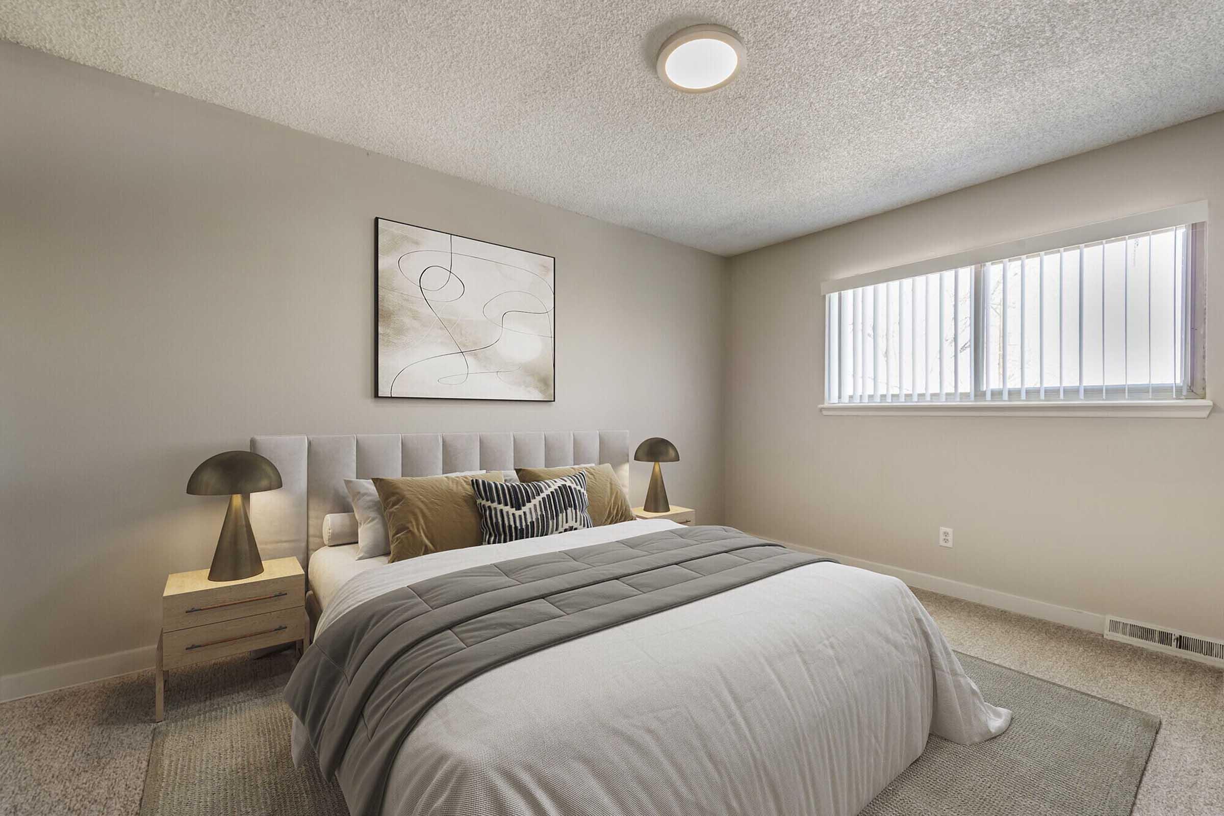 A cozy bedroom featuring a neatly made bed with a light gray comforter and decorative pillows. There are two modern lamps on either side of the bed, and a minimalist artwork hangs above the headboard. The walls are a soft neutral color, and a window provides natural light.