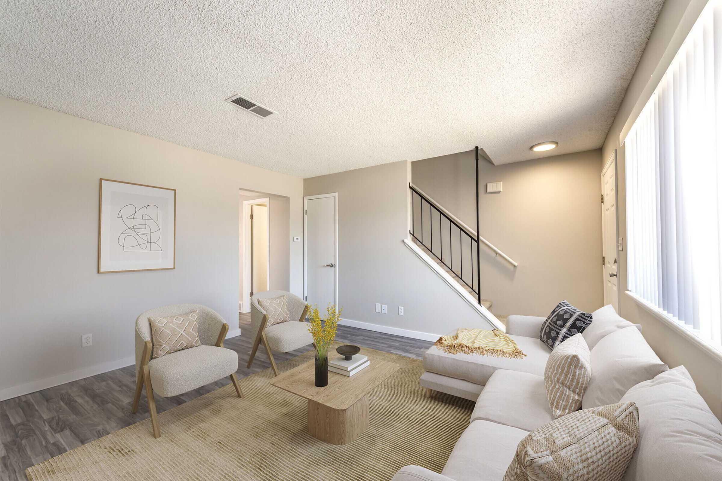 A cozy living room featuring a light-colored sofa and two patterned armchairs. A wooden coffee table sits on a textured rug. Natural light streams in through large windows, illuminating the neutral walls and the minimalist decor. A staircase leads to the upper level in the background, enhancing the inviting atmosphere.