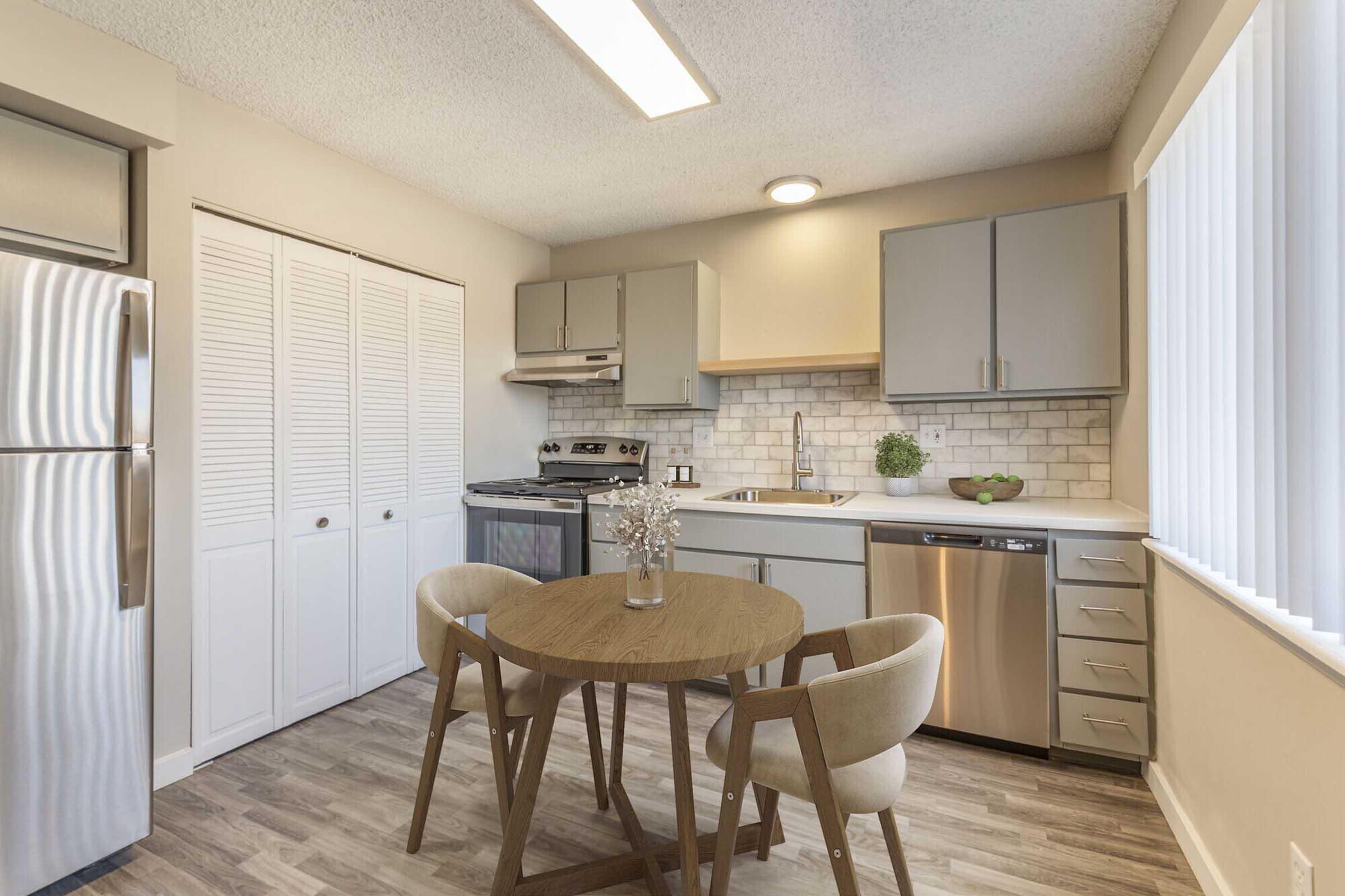 A modern kitchen featuring stainless steel appliances, including a refrigerator and oven, light gray cabinetry, and a small round dining table with two beige chairs. The space has a bright and airy feel, with natural light coming through vertical blinds and stylish tile backsplash.