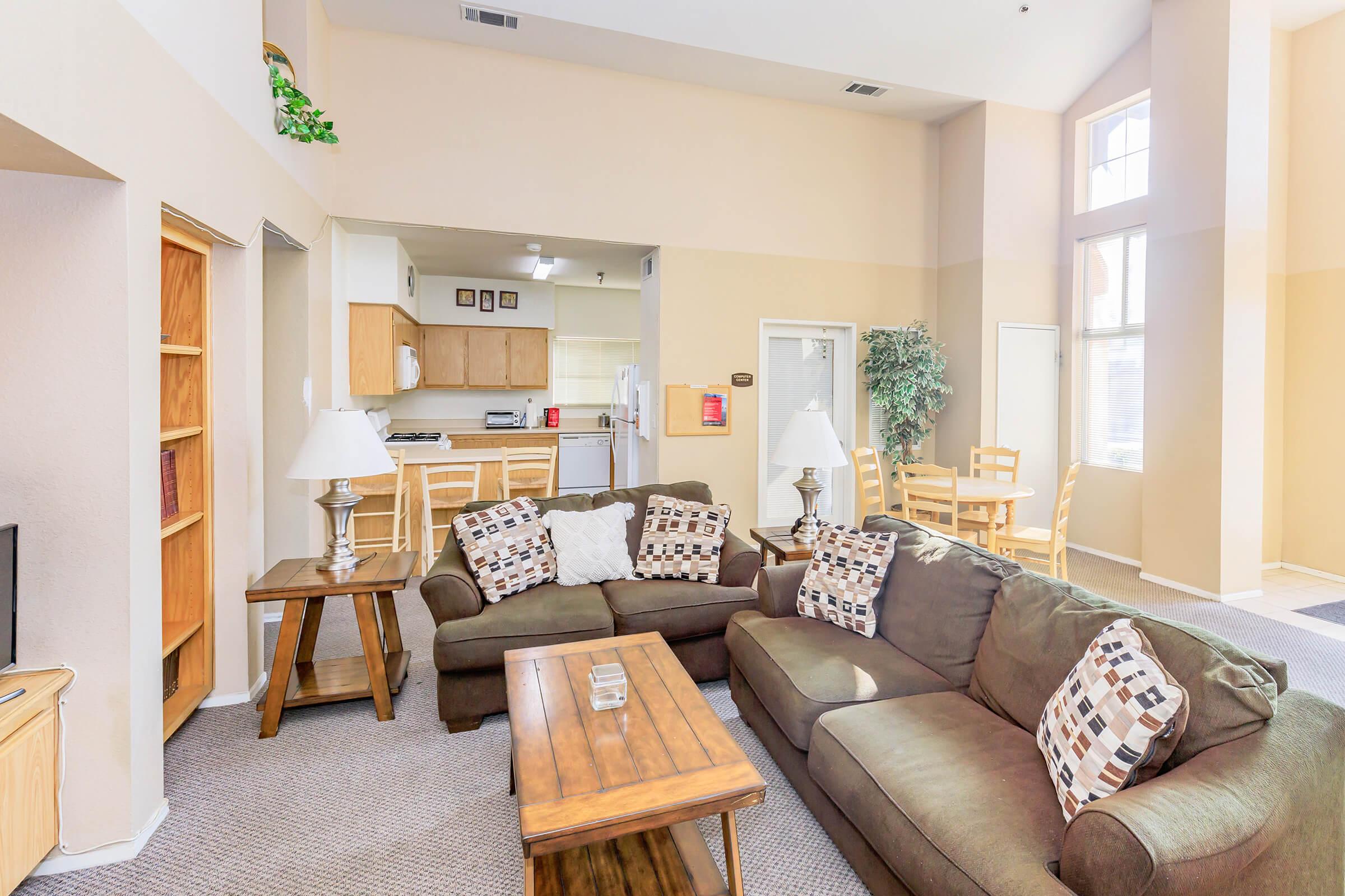 A spacious living room featuring a brown couch with decorative pillows, a wooden coffee table, and a bookshelf. The room has tall windows allowing natural light to flood in, and a kitchenette is visible in the background with wooden dining chairs and appliances. The overall decor is neutral and inviting.