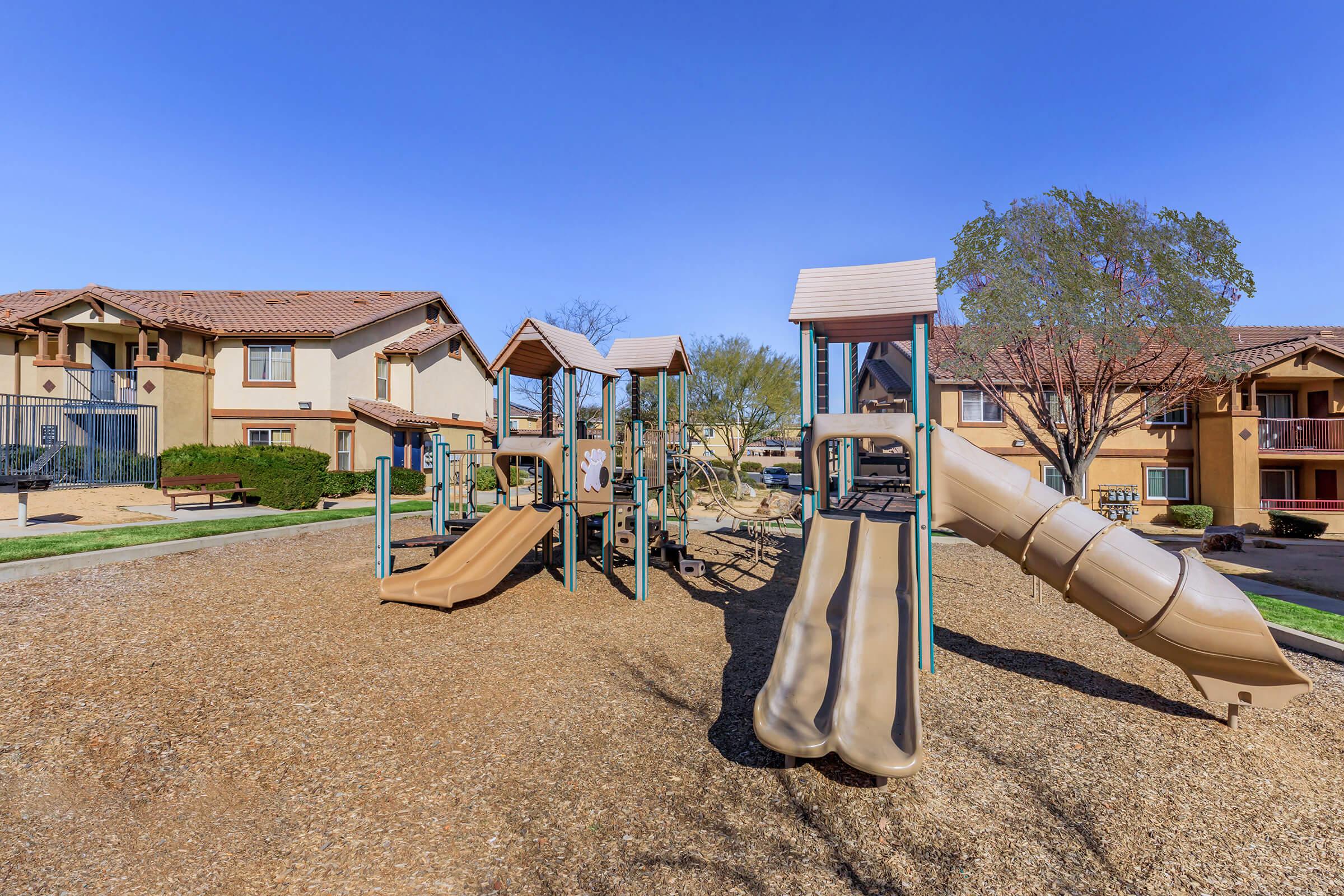 A playground with various play structures, including two slides, situated on a gravel surface. Surrounding the playground are residential buildings with a clear blue sky above. The area appears sunny and inviting, ideal for children to play.