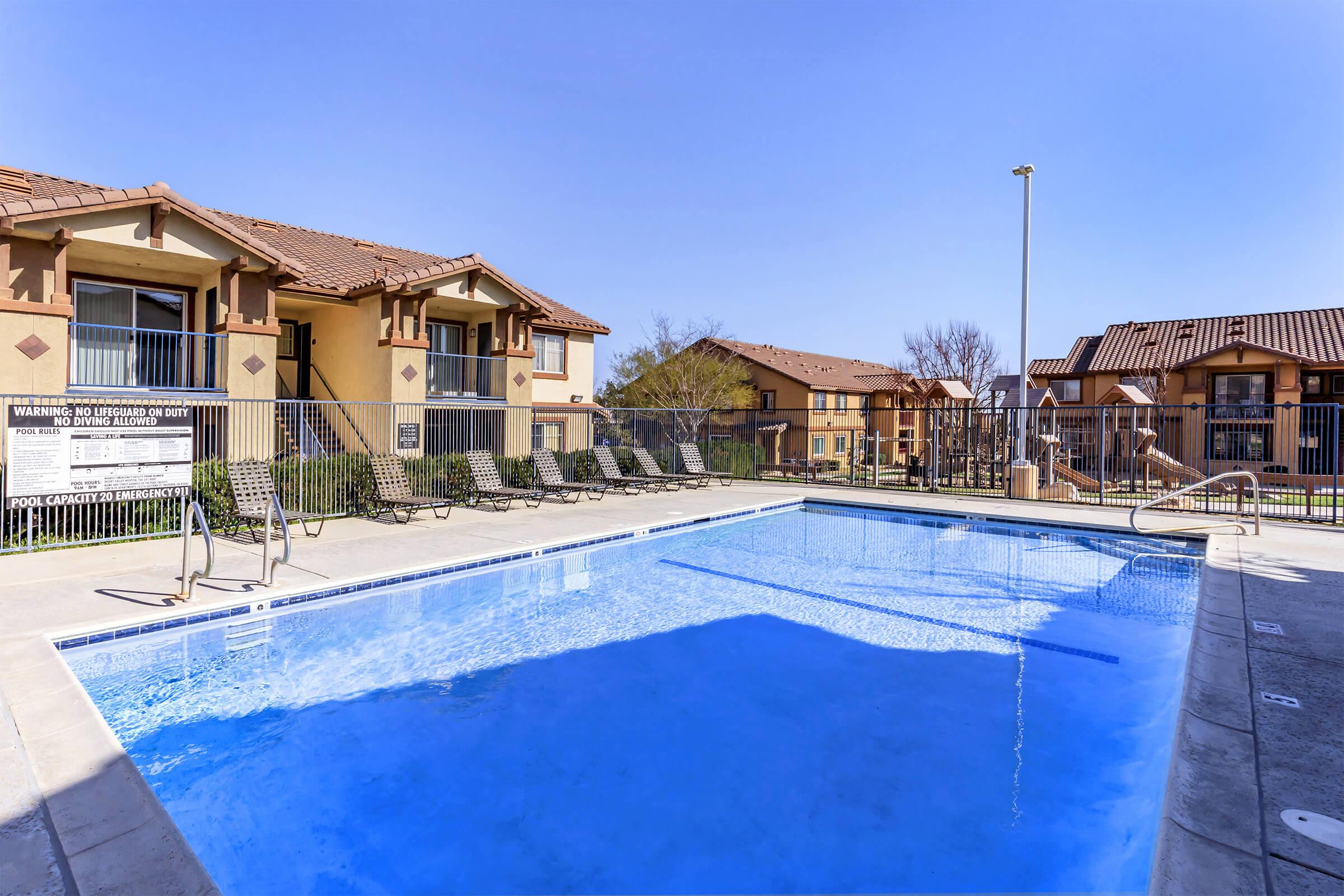 A clear blue swimming pool surrounded by lounge chairs, set against a backdrop of apartment buildings. The sky is sunny and blue, creating a warm, inviting atmosphere. A fence encloses the pool area for safety.