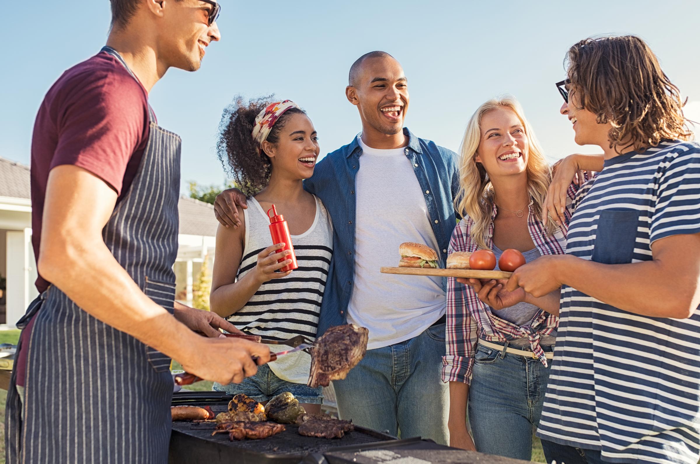 A group of five people enjoying a barbecue outdoors. One person is grilling while others engage in conversation and laughter. The scene features colorful food items, including meat and vegetables, and a relaxed, cheerful atmosphere on a sunny day.