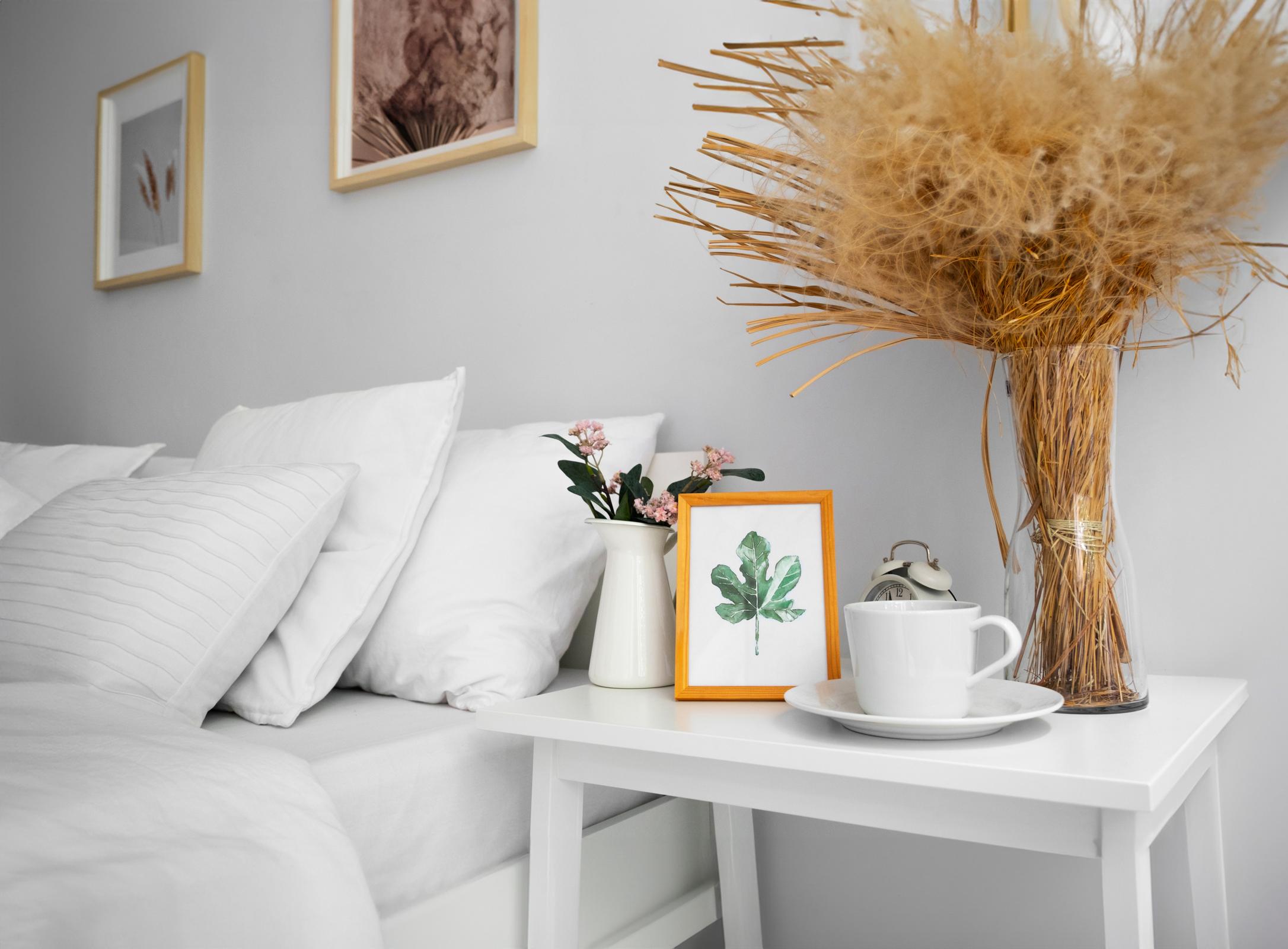 A cozy bedroom scene featuring a neatly made bed with white pillows and a small bedside table. On the table, there is a white cup, a decorative vase with flowers, and a framed watercolor leaf illustration. A tall vase filled with dried grass adds a warm touch to the minimalist decor.