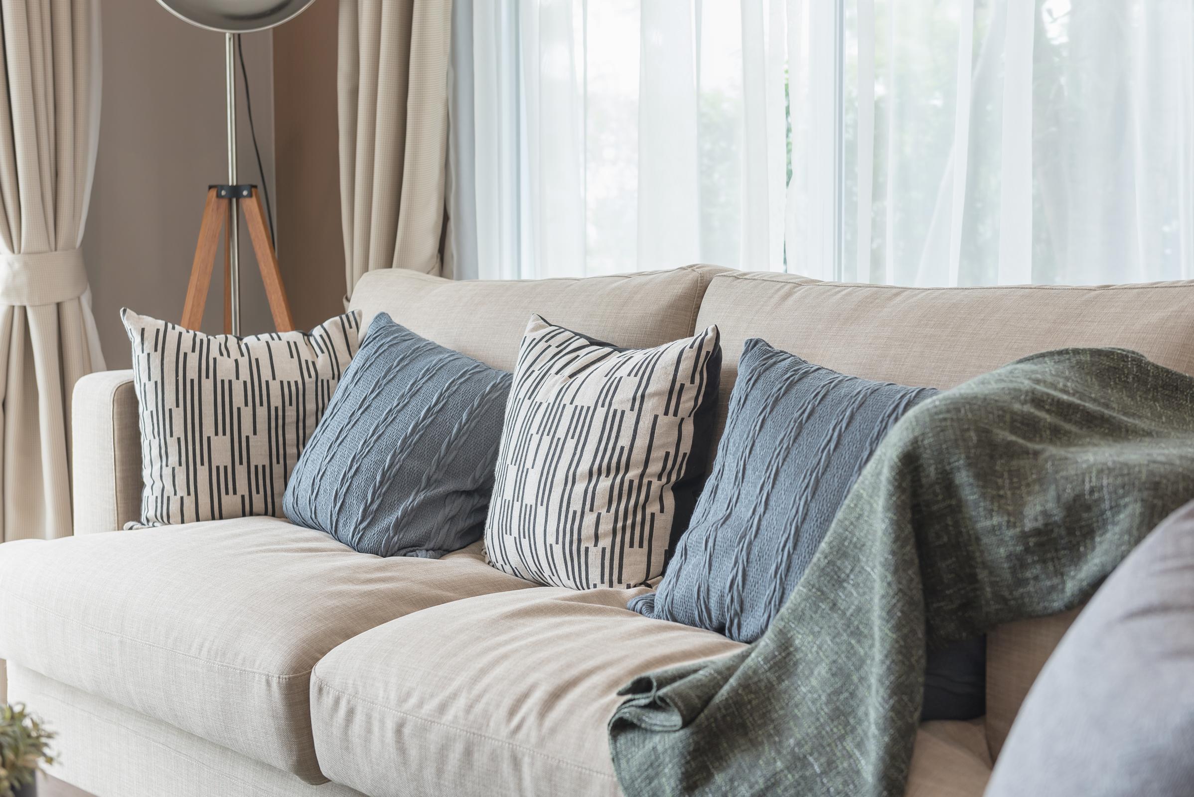 A cozy living room scene featuring a light-colored sofa adorned with patterned blue and white pillows. A textured green throw blanket is draped over one arm. Soft natural light filters through sheer curtains, creating a welcoming atmosphere.