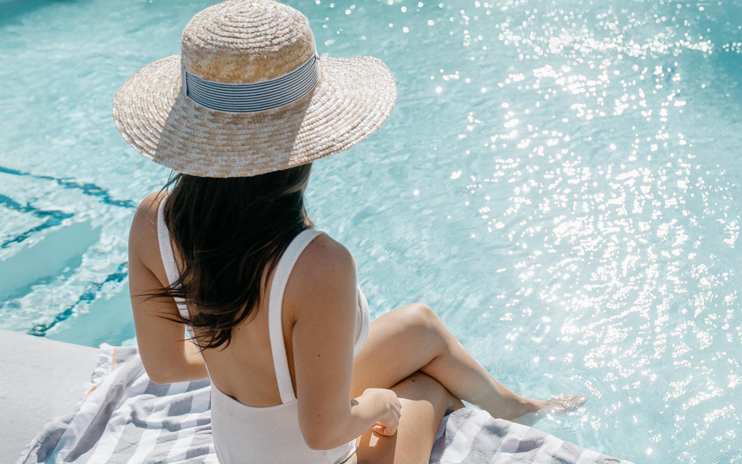 A person wearing a wide-brimmed straw hat sits by a sparkling blue pool, their back facing the camera. They are dressed in a white swimsuit and are relaxing with their legs in the water, surrounded by a striped towel. Bright sunlight reflects off the water's surface, creating a serene atmosphere.