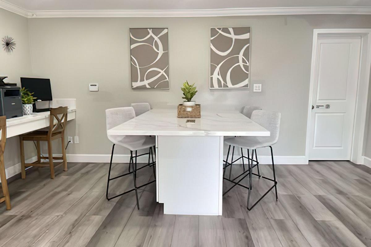 A modern kitchen/dining area featuring a large marble island with high stools, light gray walls, two abstract wall art pieces, a wooden dining table with high-backed chairs, and a doorway leading to another room. The flooring is light, resembling wood.