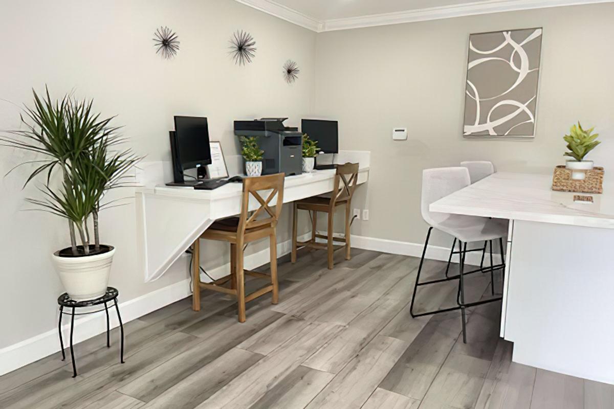 A modern workspace featuring two desks with computers and chairs, a printer, and decorative plants. Light-colored walls and wooden flooring create a bright and airy atmosphere. A large artwork piece hangs on the wall, adding a contemporary touch.