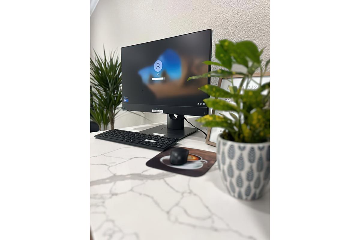 A desk setup featuring a modern computer monitor displaying a login screen, a black keyboard, a mouse on a mousepad, and a small potted plant. The surface is white with a marble pattern, and there are decorative elements in the background, including a tall green plant.