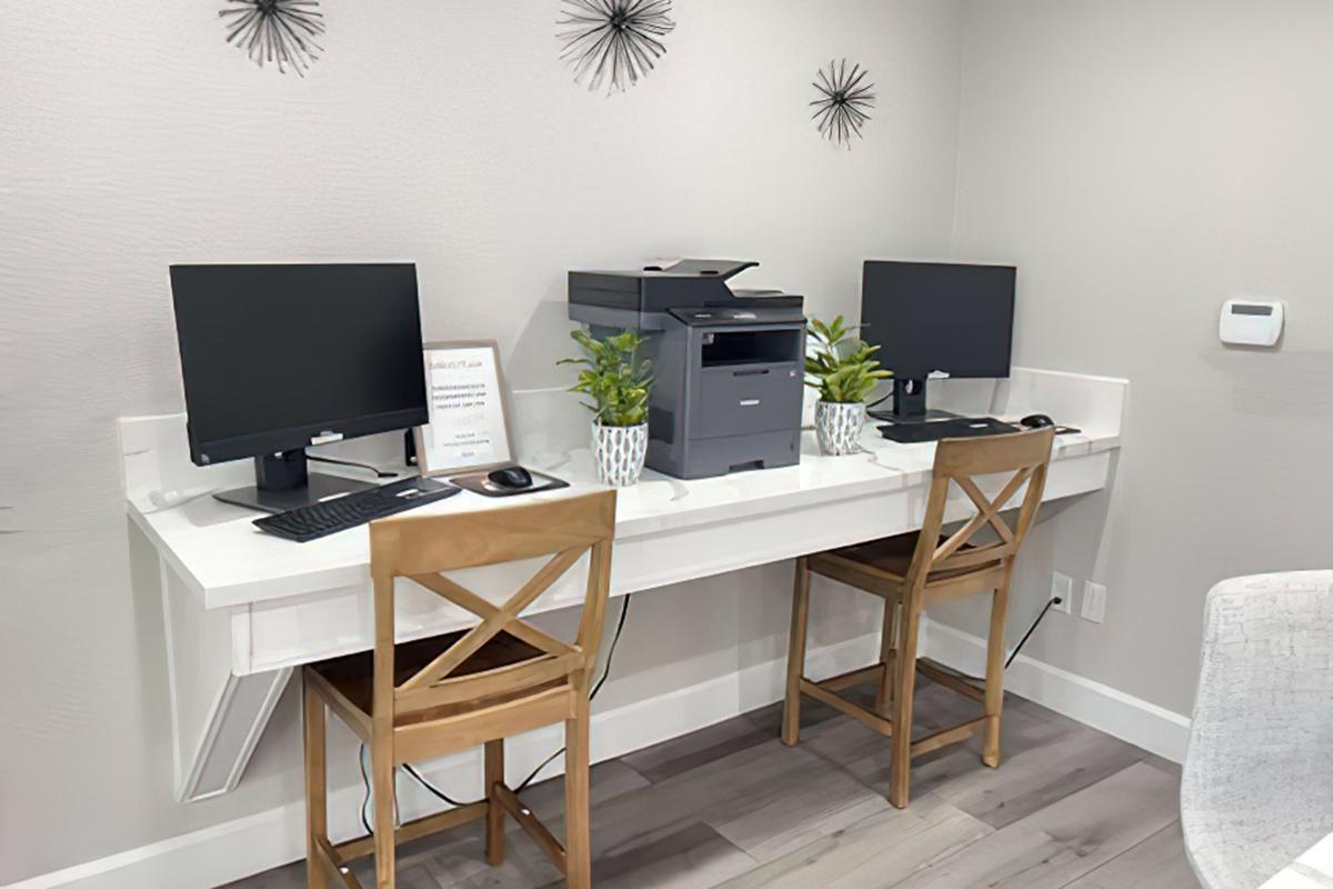 A modern shared workspace featuring two computers with monitors and keyboards placed on a sleek white desk. A printer is located on the desk, alongside two potted plants. Two wooden chairs are positioned at the desk. The wall is decorated with minimalist wall art and there's a sign on the desk.