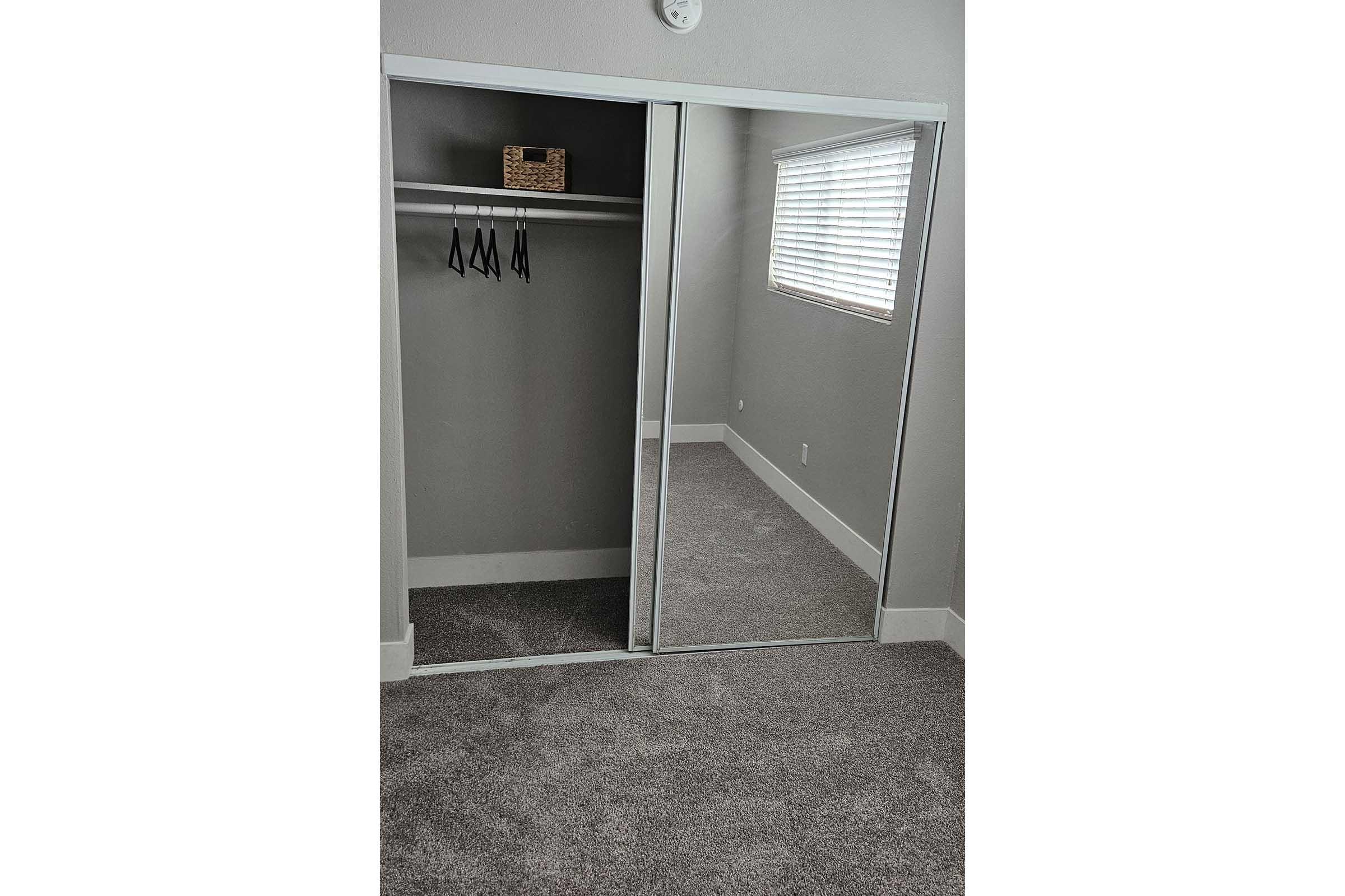 A cozy room featuring a mirrored closet with sliding doors. Inside the closet, there are several hangers and a small basket on a shelf. The floor is covered with plush, light-colored carpet, and there's a window with blinds allowing natural light to filter in. The walls are painted a soft gray.
