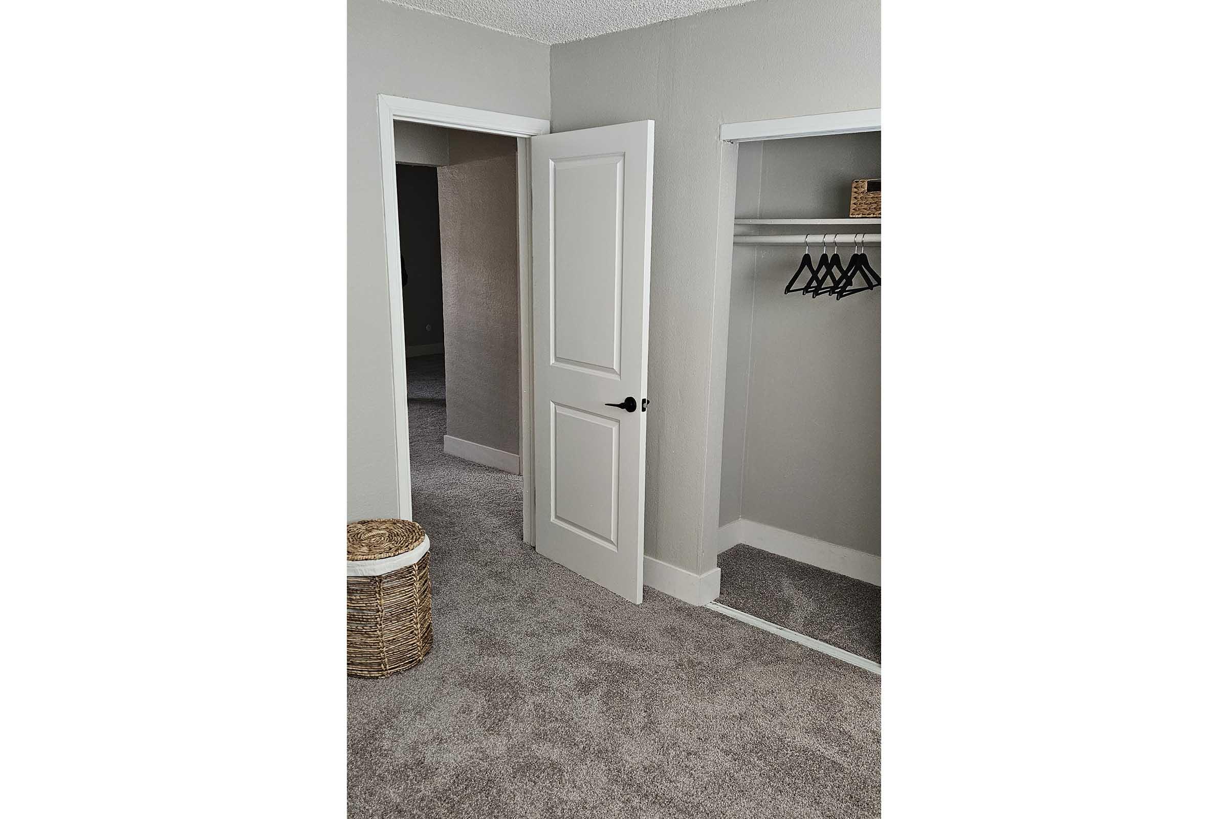A minimalistic room featuring two doorways: one leading to a closet with a hanging rod and black hangers, and the other doorway appears to lead to another room. The walls are painted light gray, and the floor is covered in soft, beige carpet. A woven basket sits in the corner.