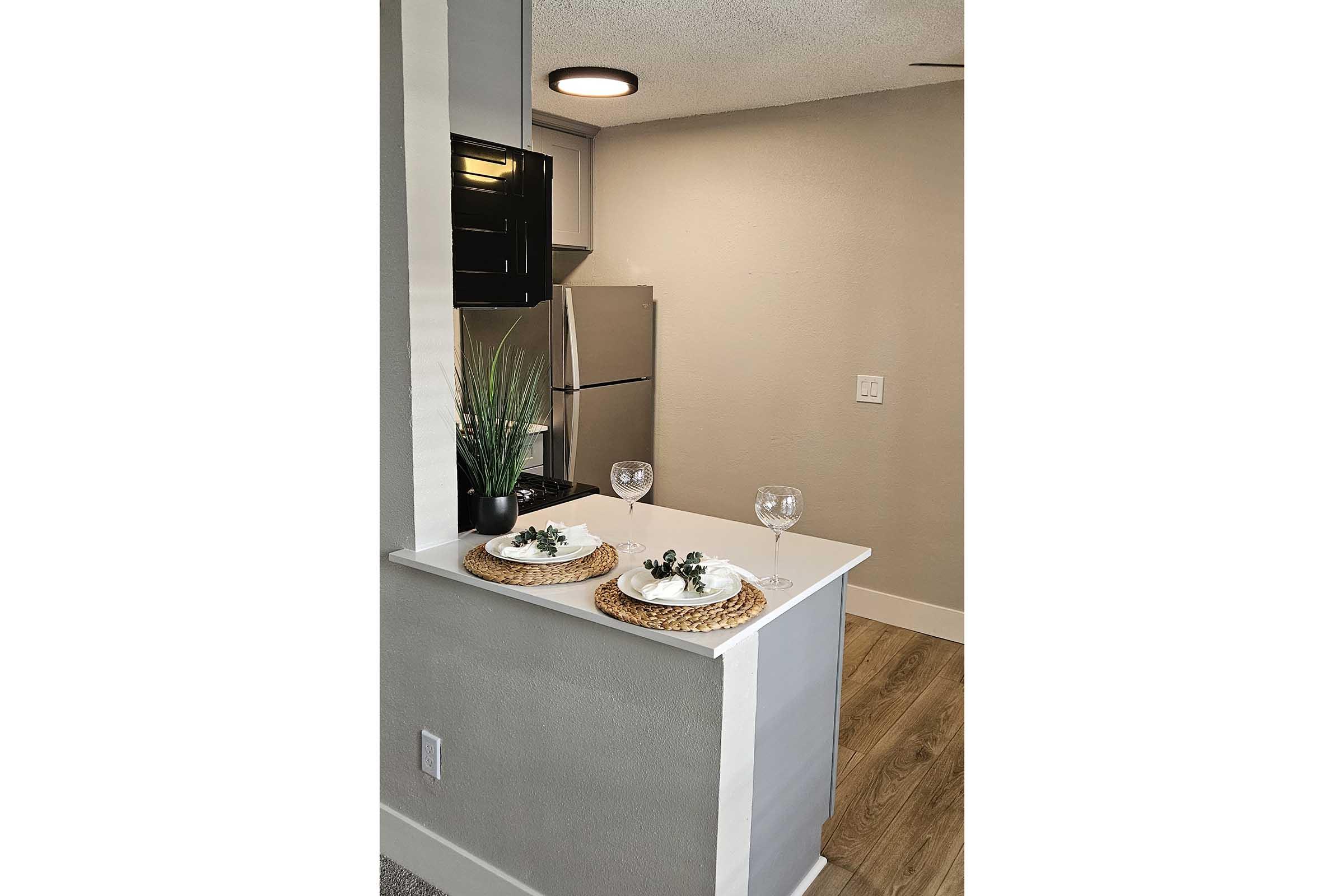 A modern kitchen nook featuring a countertop with two decorative plates, each topped with a small plant, and two wine glasses. In the background, there is a refrigerator and a neutral-colored wall. The floor has wood-like laminate, adding to the contemporary feel of the space.