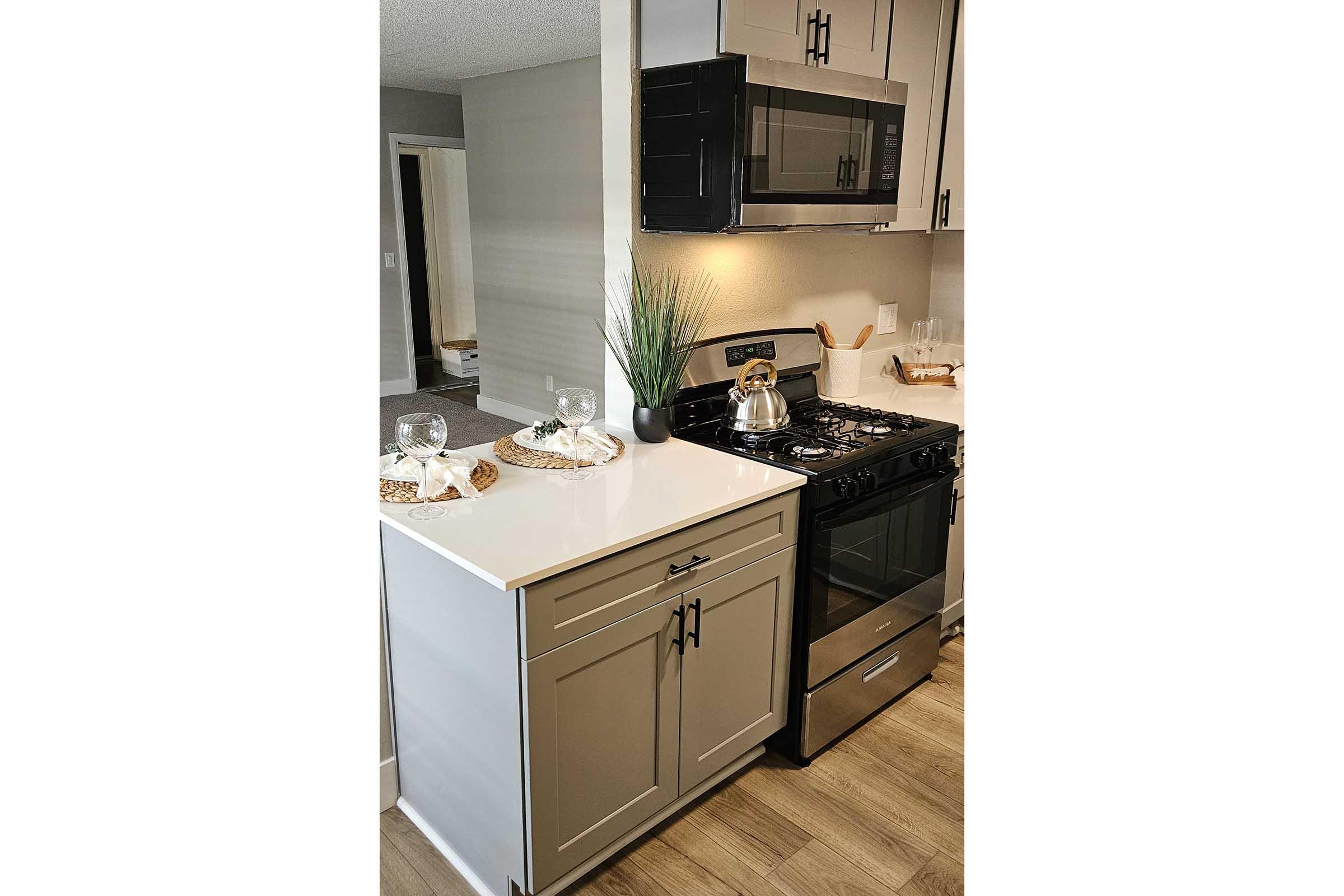 A modern kitchen featuring light-colored cabinetry, a stainless steel gas stove, and a microwave above. The countertop is white with a decorative tray, glass dish, and plant. The flooring is wooden and there is a glimpse of adjacent living space in the background.