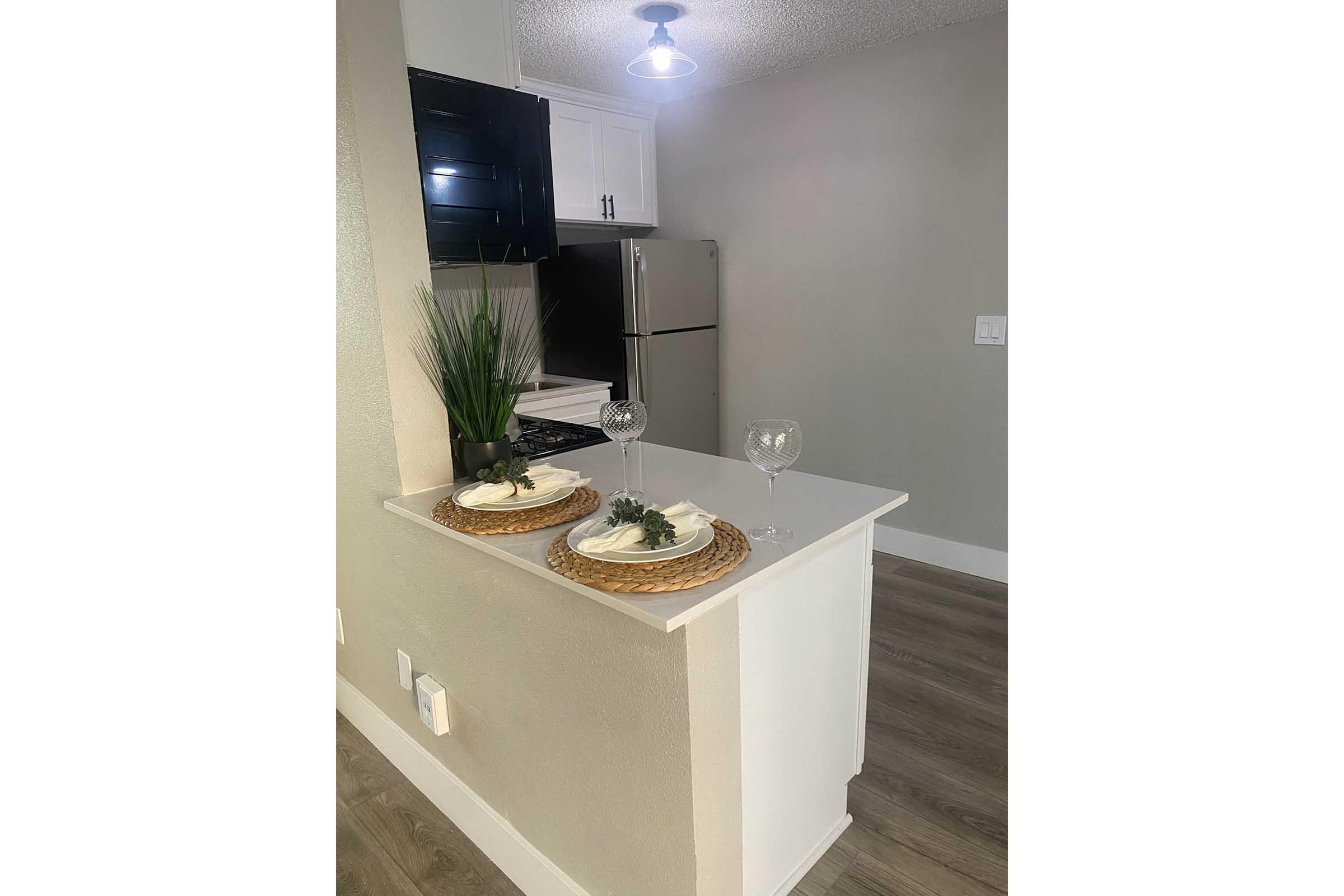 A modern kitchen space featuring a white countertop with two decorative plates topped with plants, alongside two wine glasses. The kitchen has black cabinetry, stainless steel appliances, and a stylish light fixture above. The flooring is a light wood color, creating a bright and inviting atmosphere.